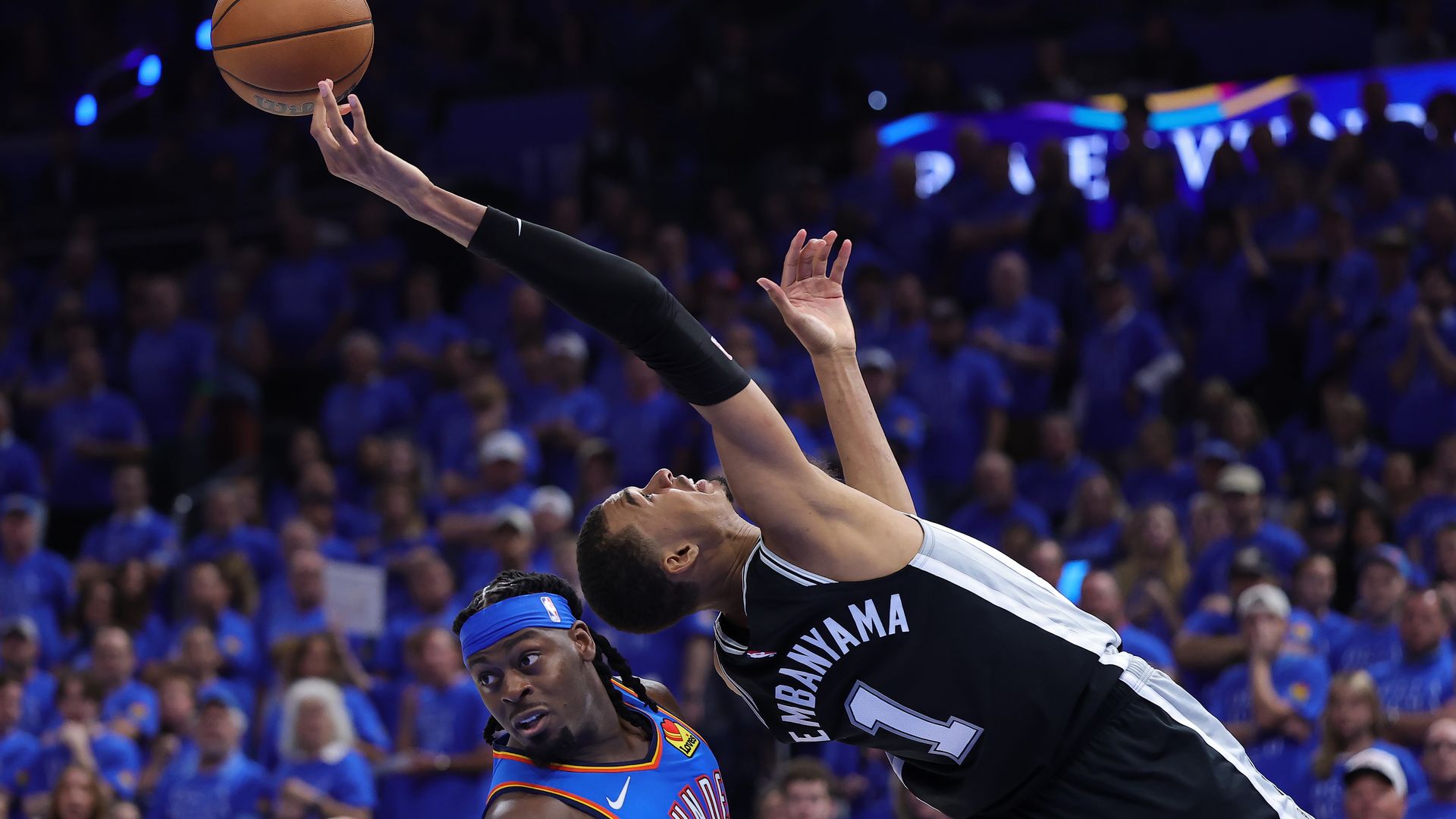 The Spurs' Victor Wembanyama is leaning backward over the Thunder's Luguentz Dort. Wemby is reaching for a basketball with his fingertips and Dort is looking to the sidelines. A sea of fans in blue shirts is blurred in the background in Game 1 of the NBA Western Conference Finals in Oklahoma City.
