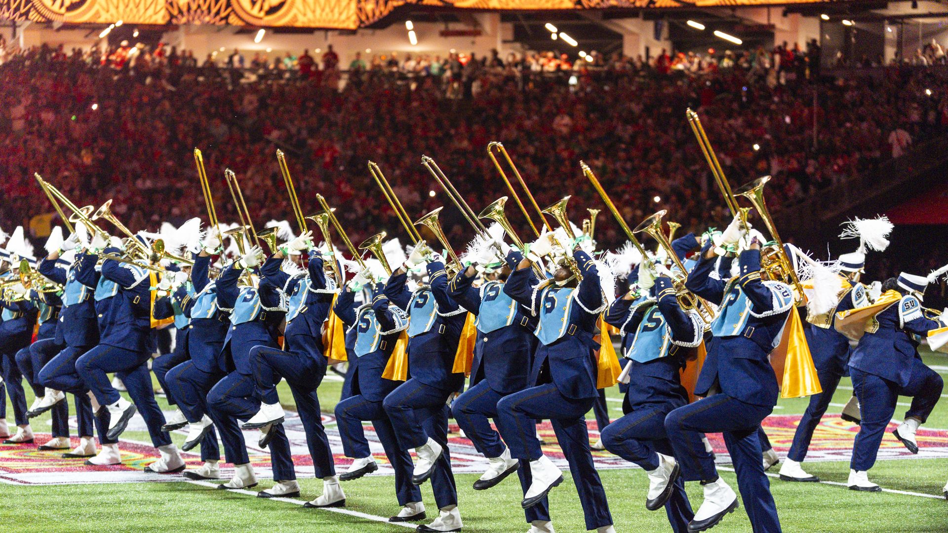 Marching band musicians blow on trombones as they all march in a line on a football field.