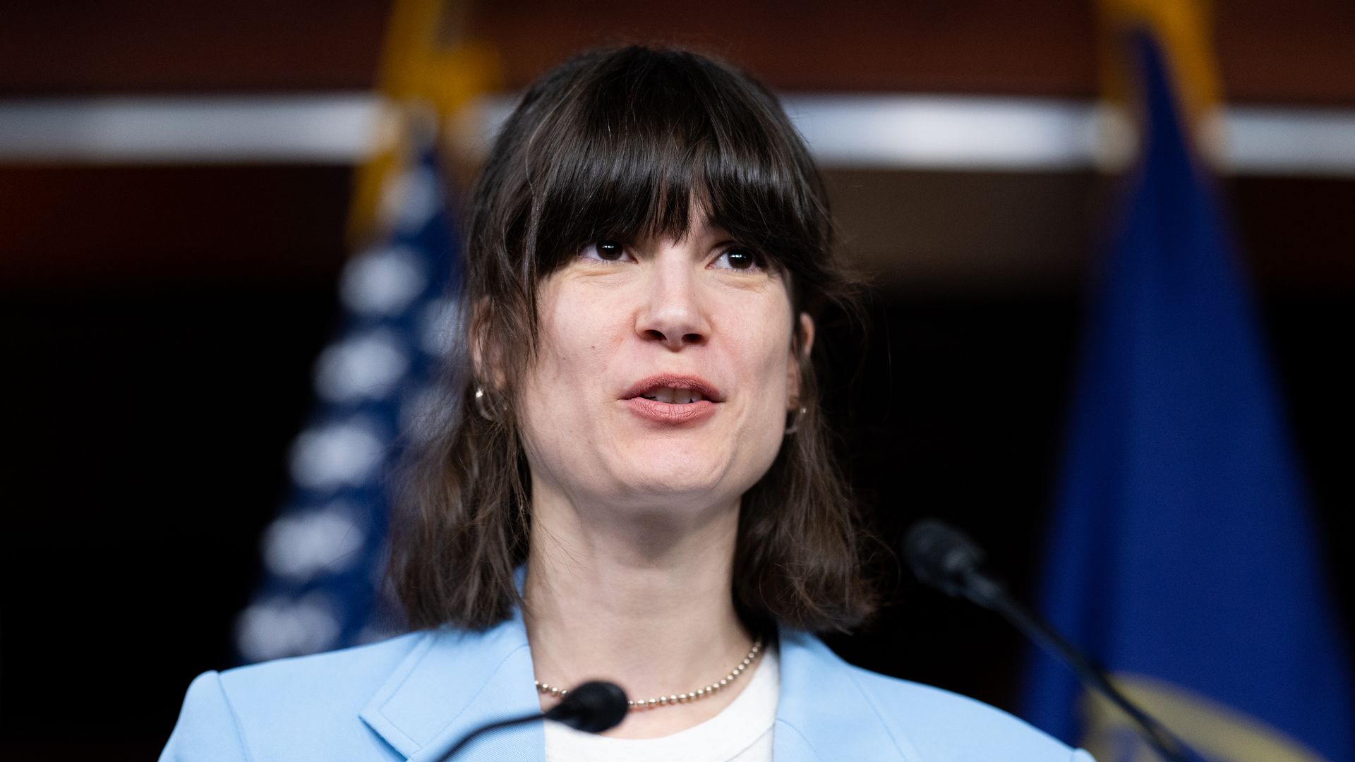 Rep. Marie Gluesenkamp Perez, wearing a powder blue blazer and white sblouse, speaking at a microphone in front of blue flags.