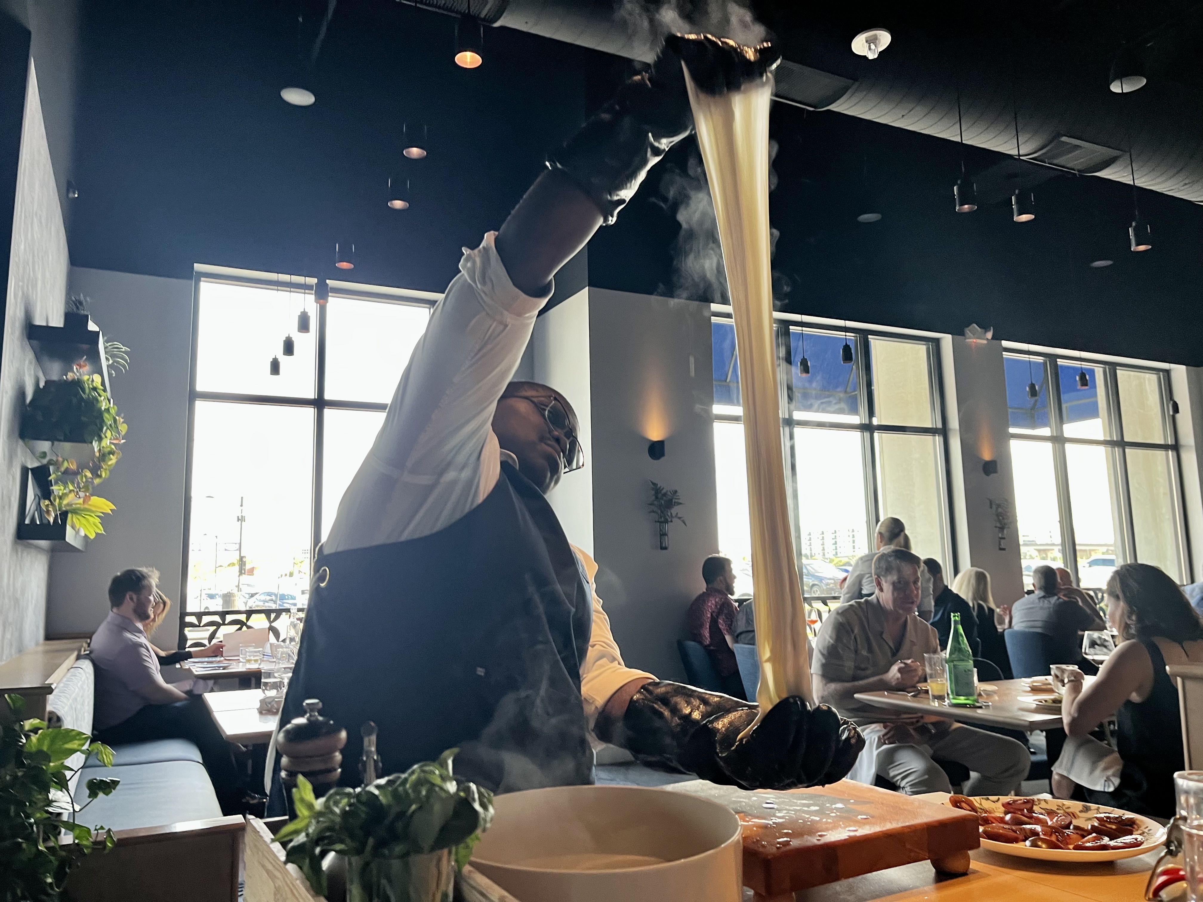 Busy restaurant interior with diners by large windows. A server in a white shirt and black apron raises an arm to stretch a giant piece of white cheese.