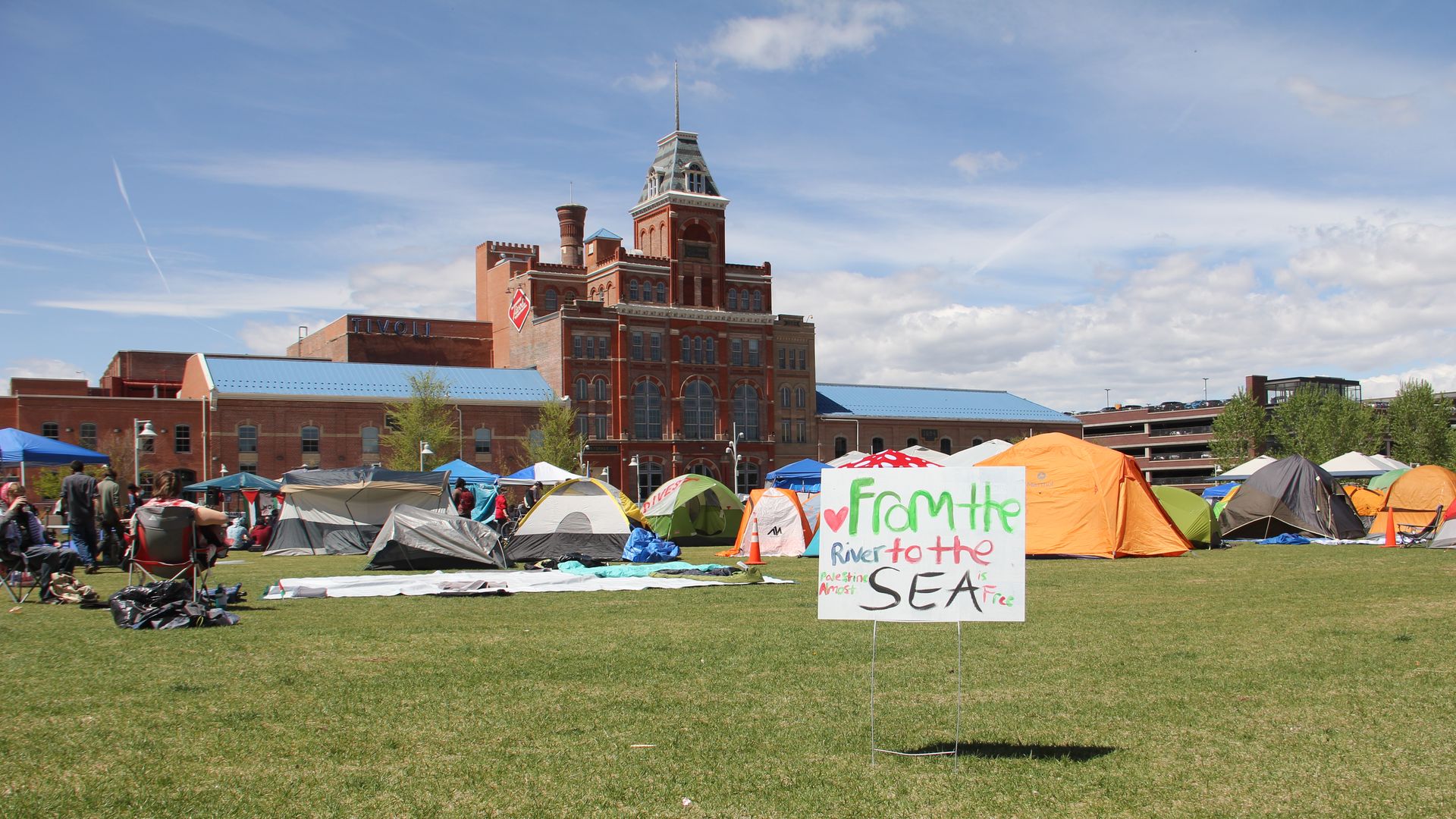 A sign in the foreground reads FROM THE RIVER TO THE SEA planted on a lawn, while several camping tents of various colors are visible in the background. In the far background, a large brick and historic building is visible during a sunny day. 