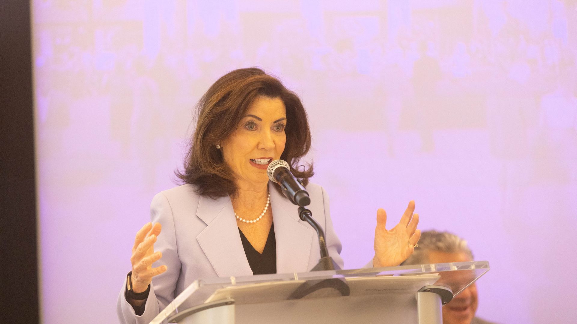 New York Gov. Kathy Hochul, wearing a lilac jacket, black top and white pearls, gestures with both hands as she speaks into a mic before a pink background.