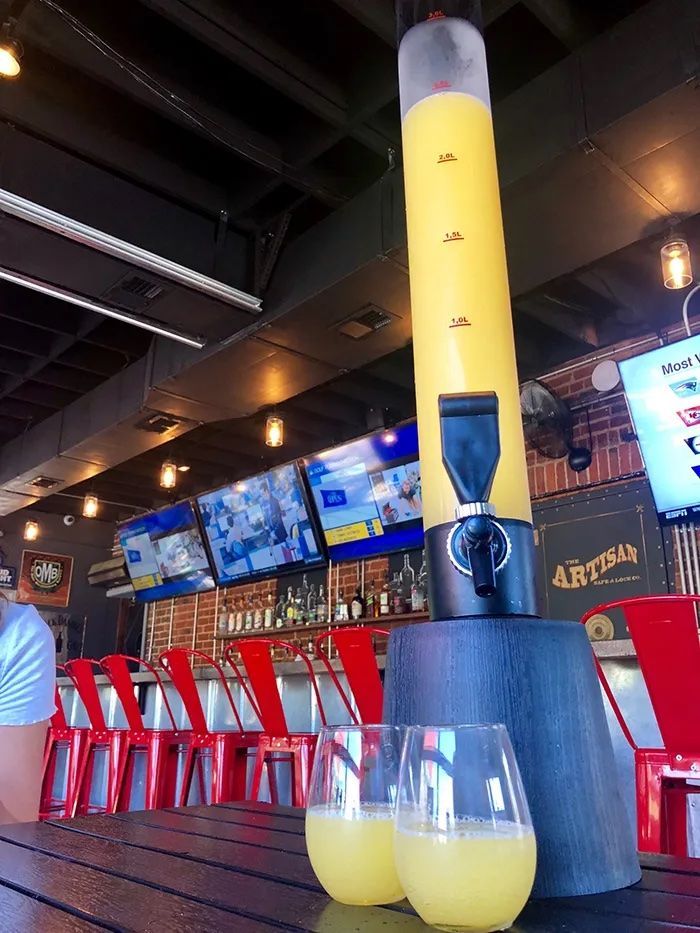 Indoor bar with a tall yellow beer-tap tower on a blue base, marked with ml lines. Red stools line brick bar; two pale yellow drinks sit on a dark wooden table in the foreground, with TVs overhead.