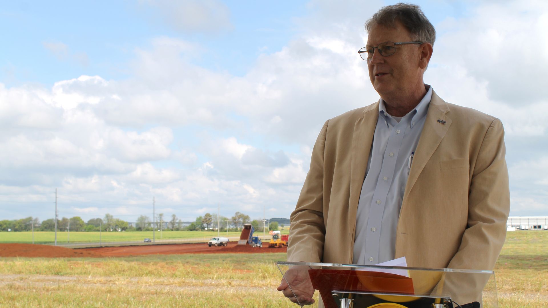 Mid-aged man in a beige blazer and light blue shirt, wearing glasses, stands at a transparent podium with a yellow hard hat, outdoors in a grassy area under a blue sky with clouds.