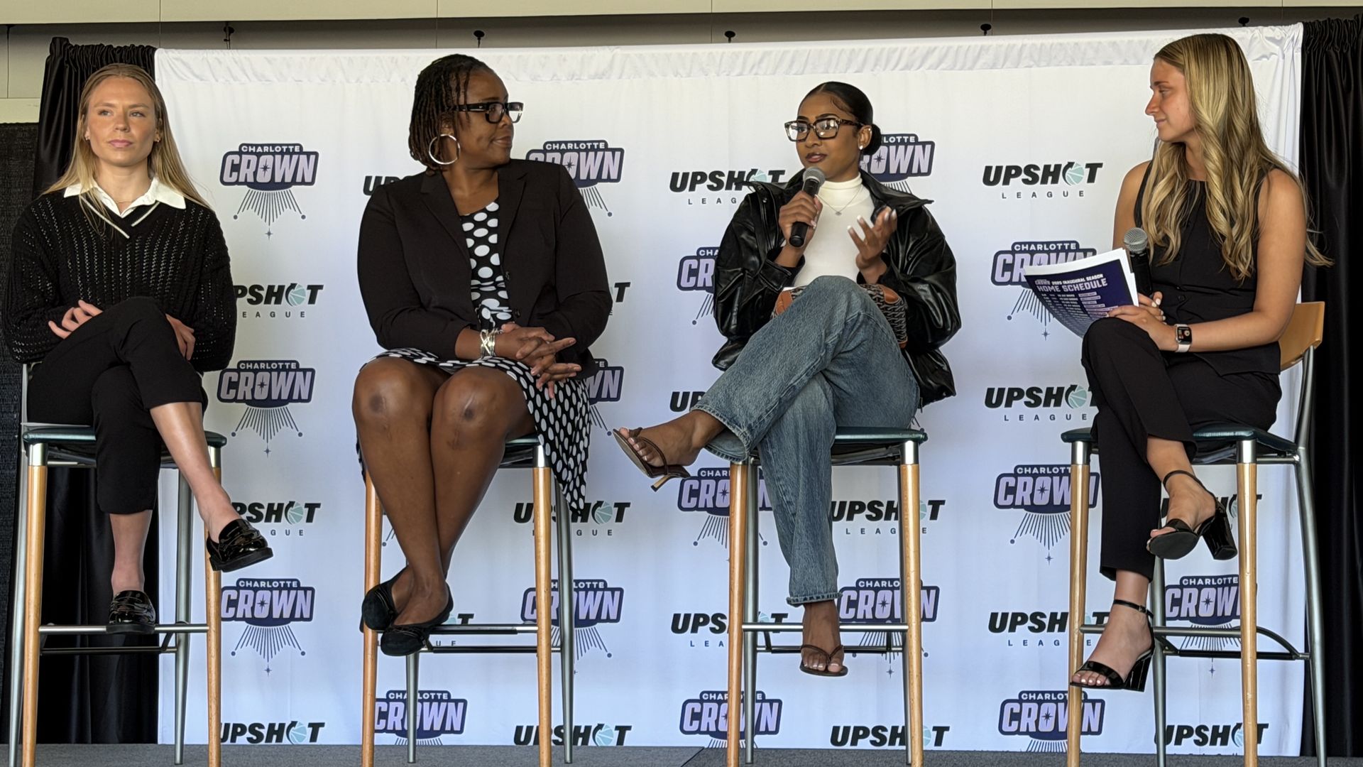 Four women panelists sit on high stools in front of a Charlotte Crown and Upshot League backdrop; the third speaker speaks into a microphone while others listen.