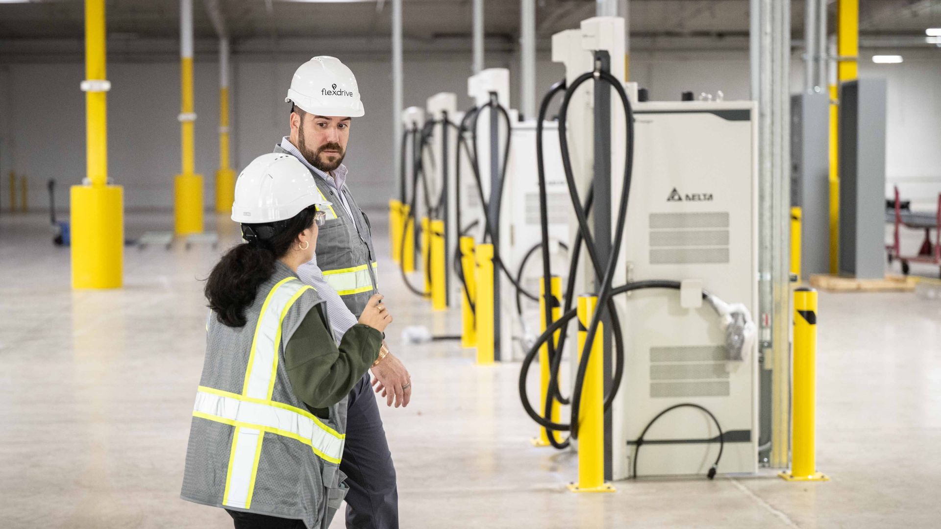 Two workers in white hard hats and high-visibility vests walk through a spacious industrial facility with yellow safety posts and large white machines with tangled black hoses.