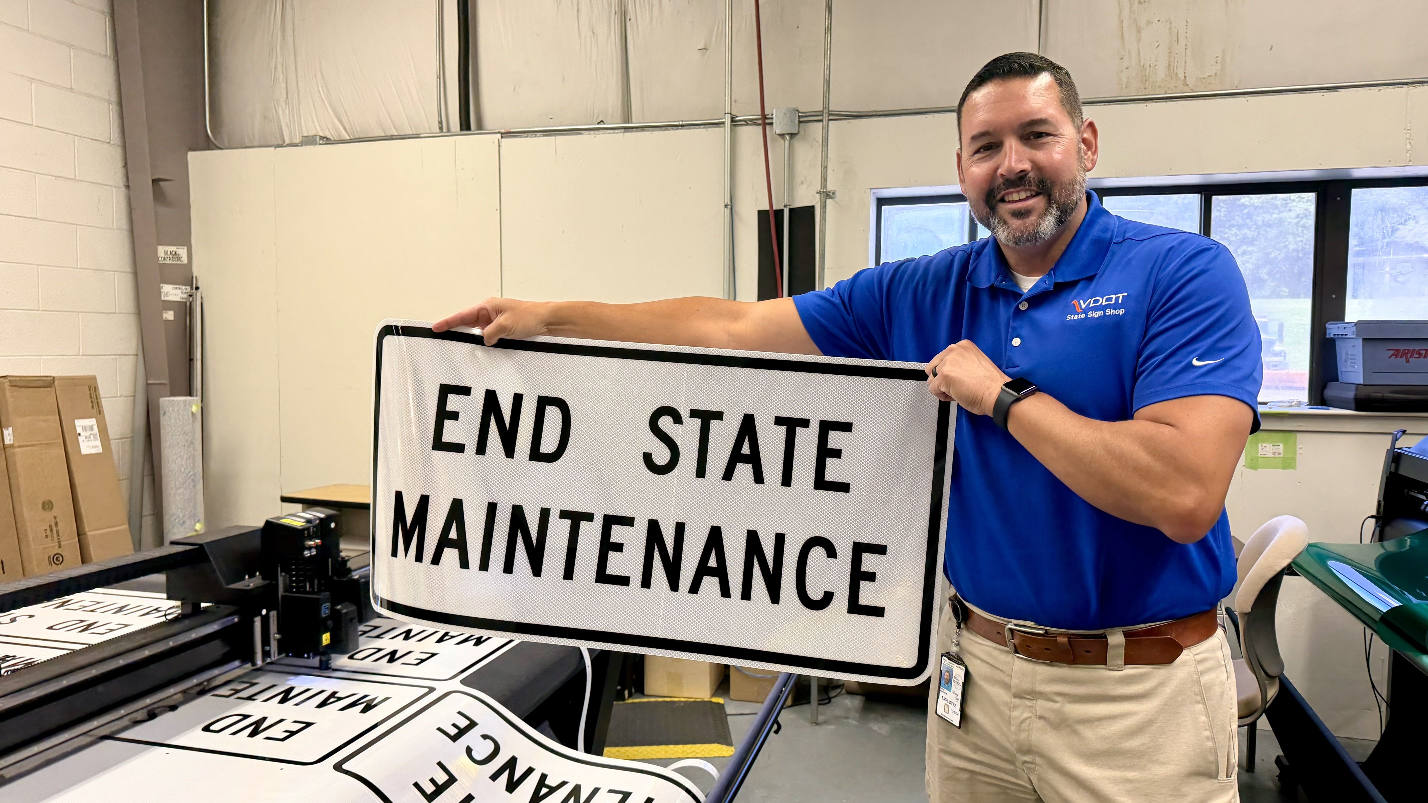 Smiling man in a blue polo holds a large white road sign with bold black text reading "END STATE MAINTENANCE" in a workshop with sign-making equipment and materials nearby.