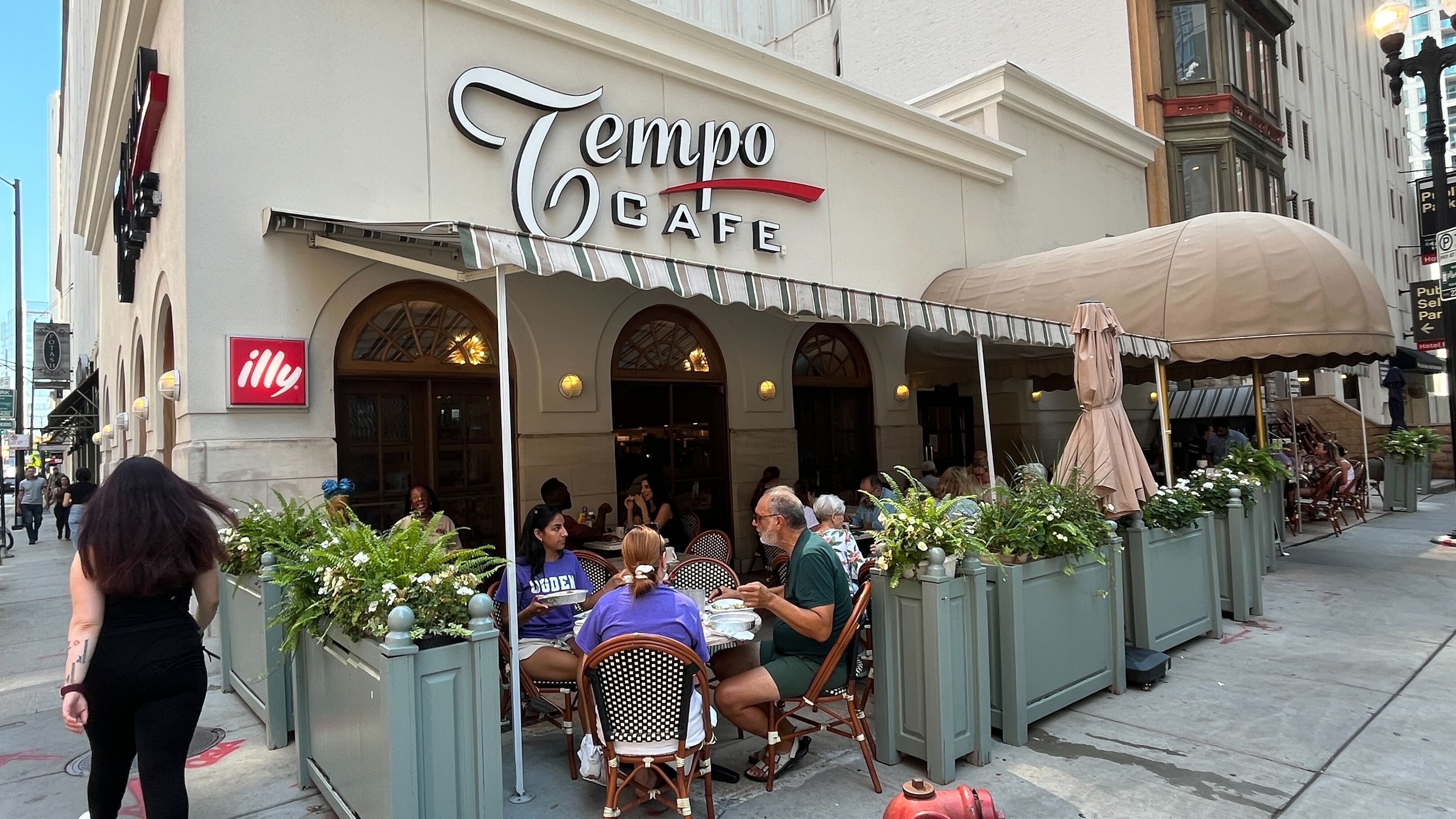 Outdoor seating at Tempo Cafe with people dining under striped awning and large beige canopy, green planters, and illy coffee sign on beige building in urban setting.