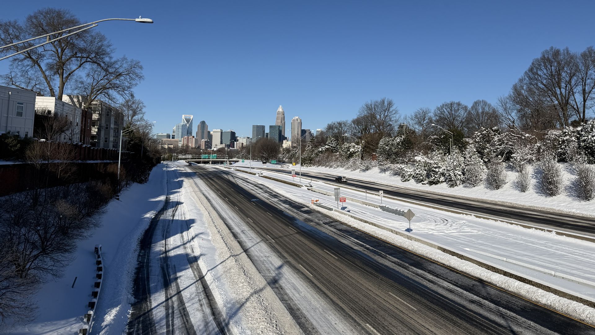 Empty snow-covered highway leading to a city skyline under a clear blue sky, with leafless trees and buildings on either side.