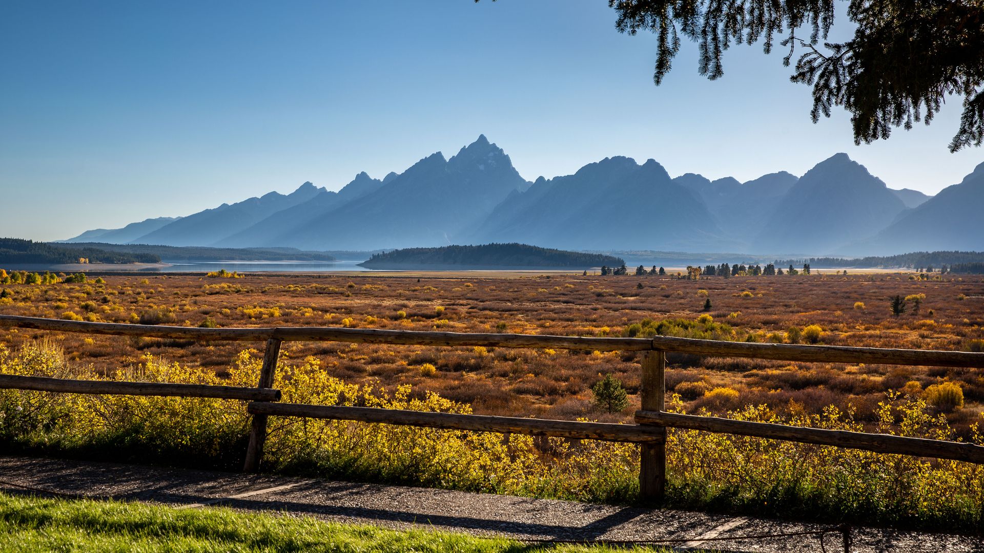 A mountain range in the background, a ranch with some cattle in the distance in the mid-range and a wooden fence in the foreground.
