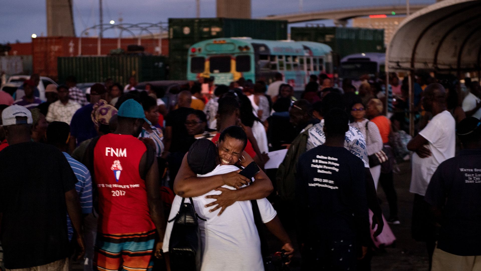 In this image, two people embrace while standing in front of a crowd of people. A bus is visible in the background.