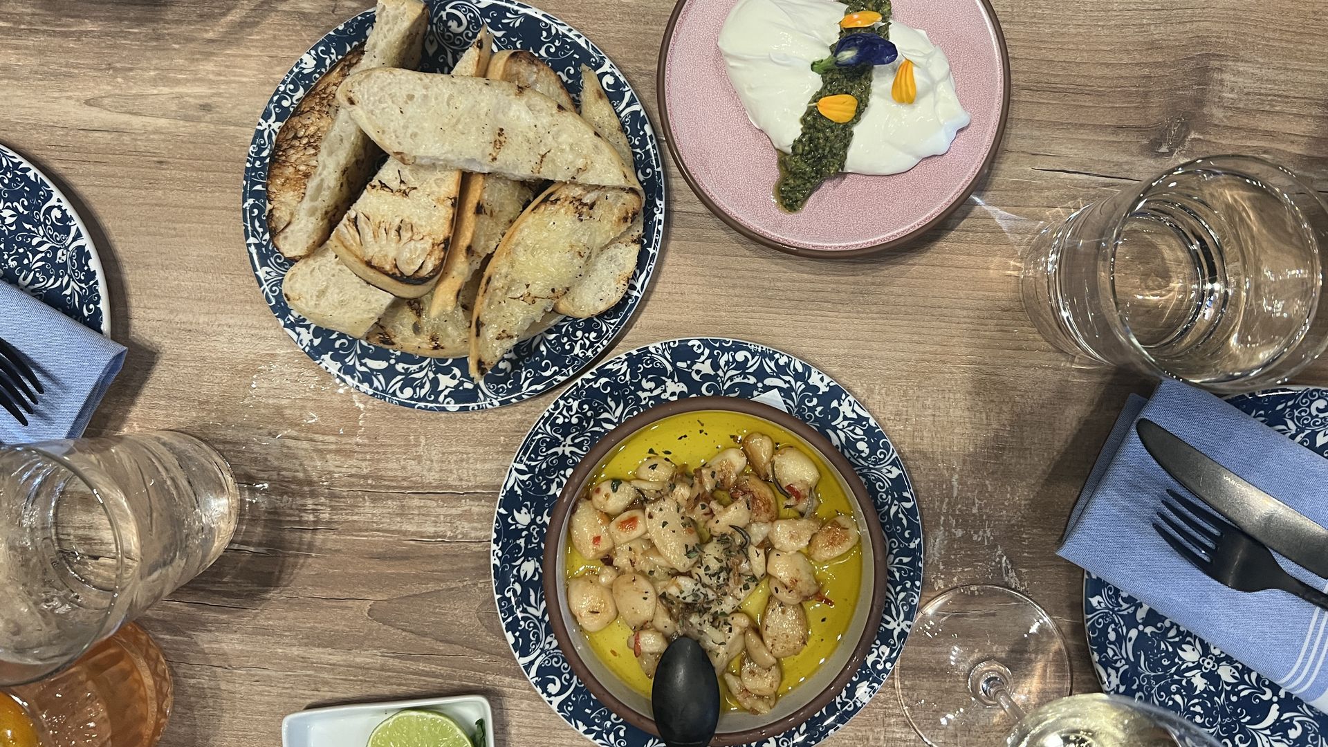 Overhead shot of a rustic table: blue patterned plates, a bread basket, pink plate with soft cheese and a green pesto stripe adorned with petals, and a yellow chickpea dish with lime wedges.