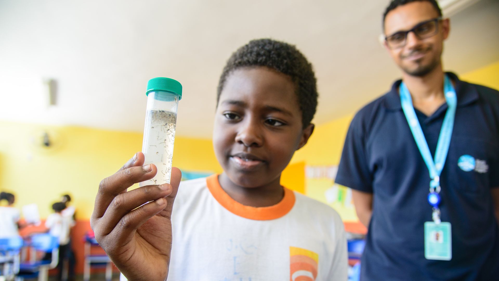 A young boy holds up a tube with mosquitos in it. A person with a lanyard stands behind him. 