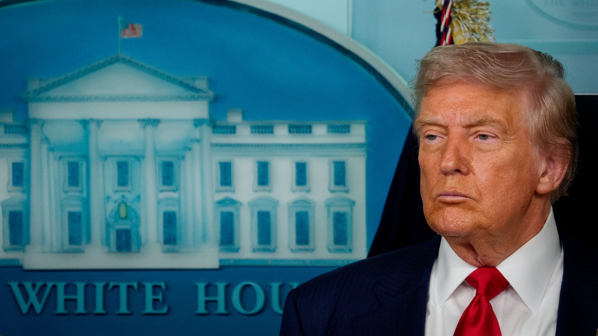 Portrait of a man resembling former President Donald Trump in a dark suit with a red tie, standing in front of a backdrop featuring the White House seal.