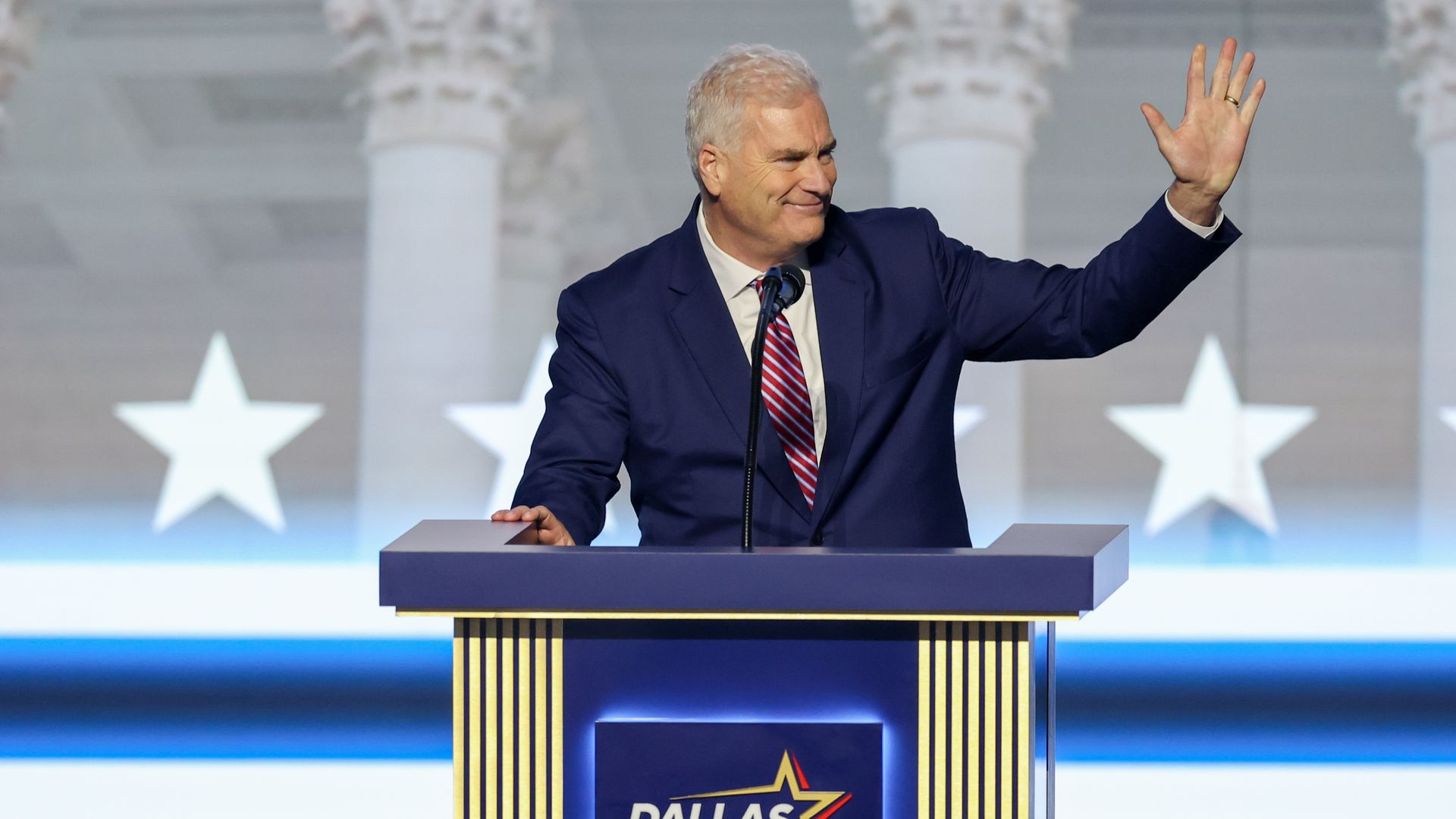 A smiling man in a dark blue suit and red striped tie stands at a podium, right hand raised in a wave. Behind him are tall columns with star shapes and a Dallas Host Committee 2026 sign.