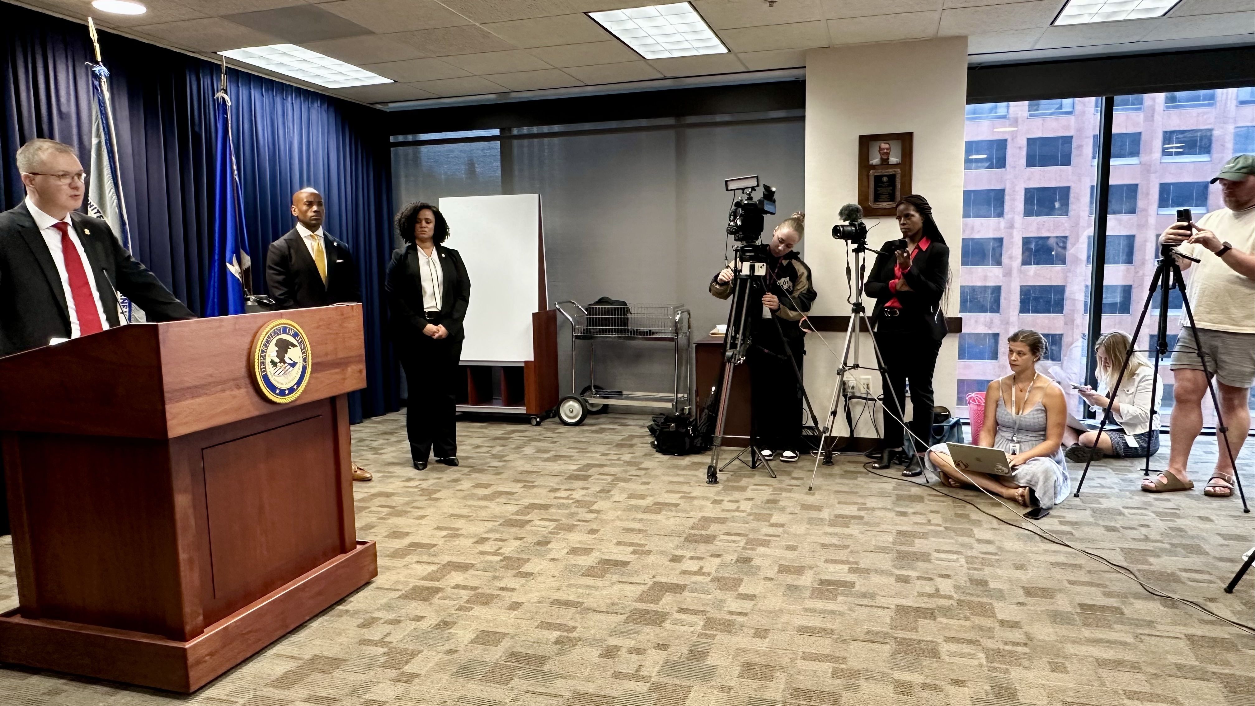 A man in a black suit and red tie speaks at a wooden podium with the Department of Justice seal, flanked by two people in business attire, while a group films and takes notes in a conference room.
