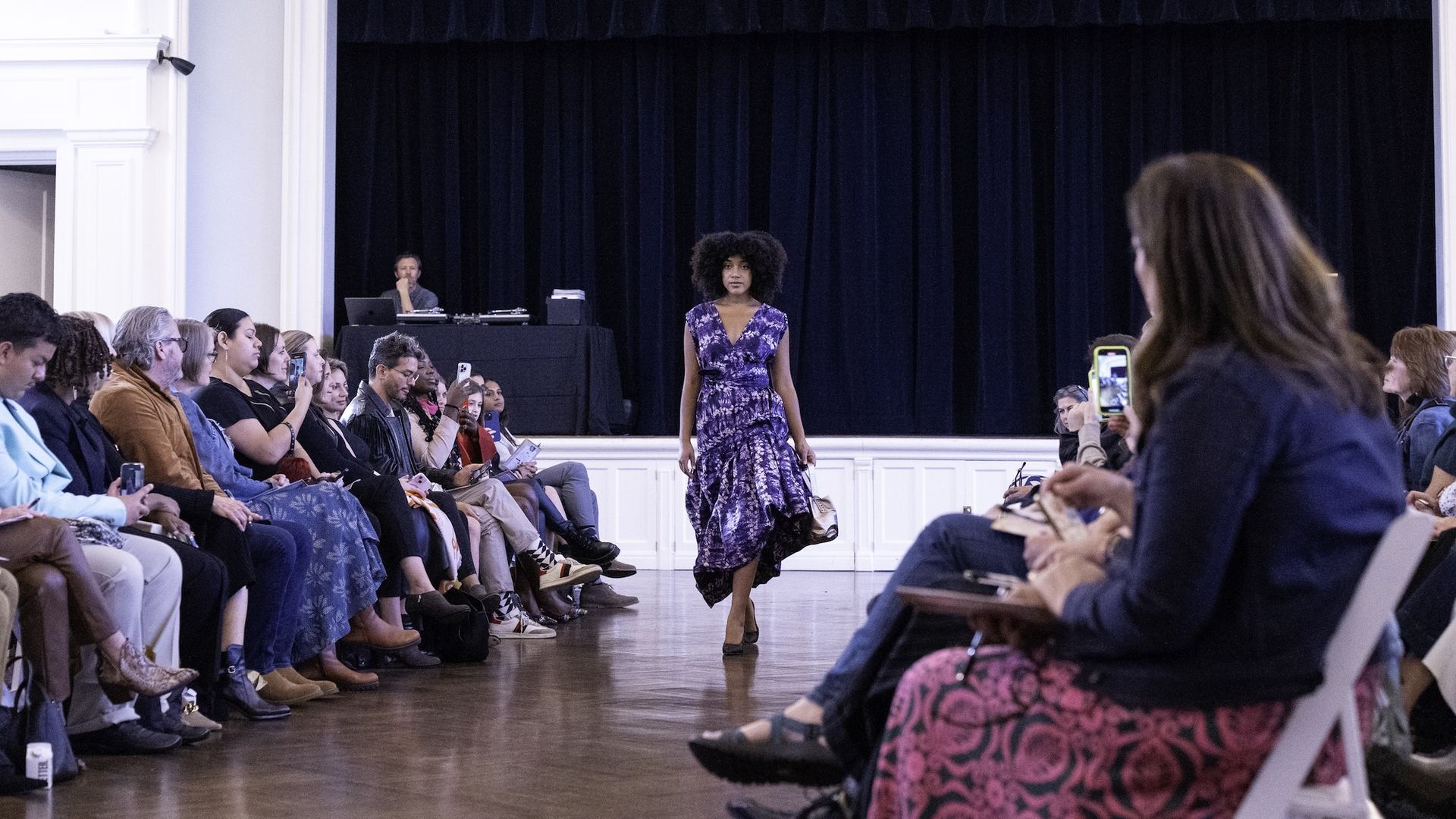A fashion model wearing a purple floral dress walks down a runway in a bright theater. Audience sits on both sides, many filming with phones, dark stage curtains behind, DJ booth to the left.