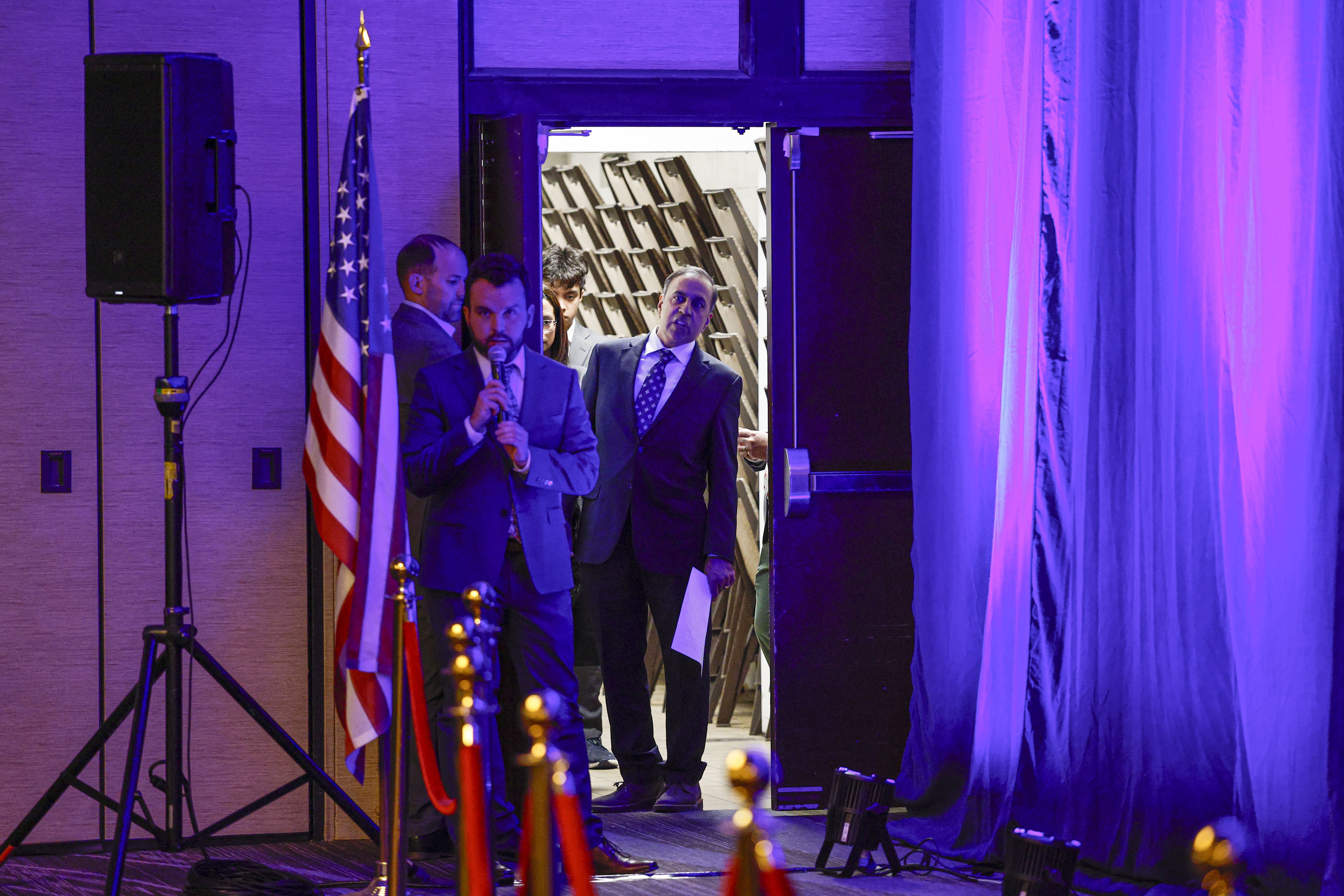 Man in a blue suit speaks into a microphone beside the American flag at a formal event; other suited attendees stand in a doorway as purple stage lights illuminate the scene.