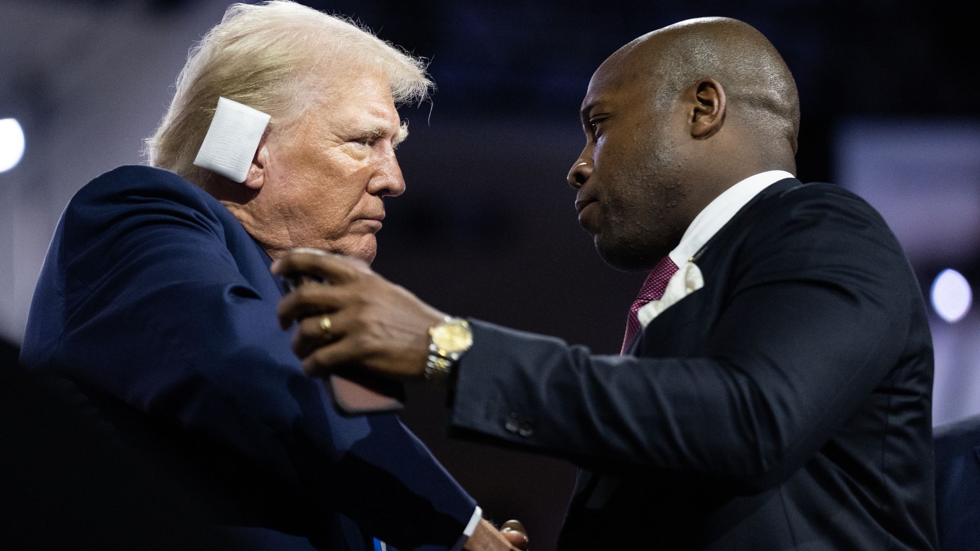 Two men in suits shaking hands and facing each other; one man with white hair and a pad on his ear, the other bald with a gold watch, in a dimly lit setting.