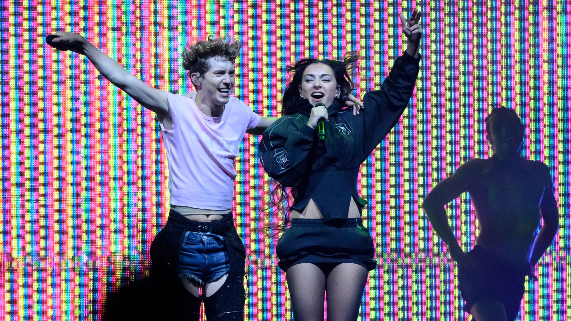 Two individuals performing on stage jump and smile in front of a multicolored screen as a person dances behind them