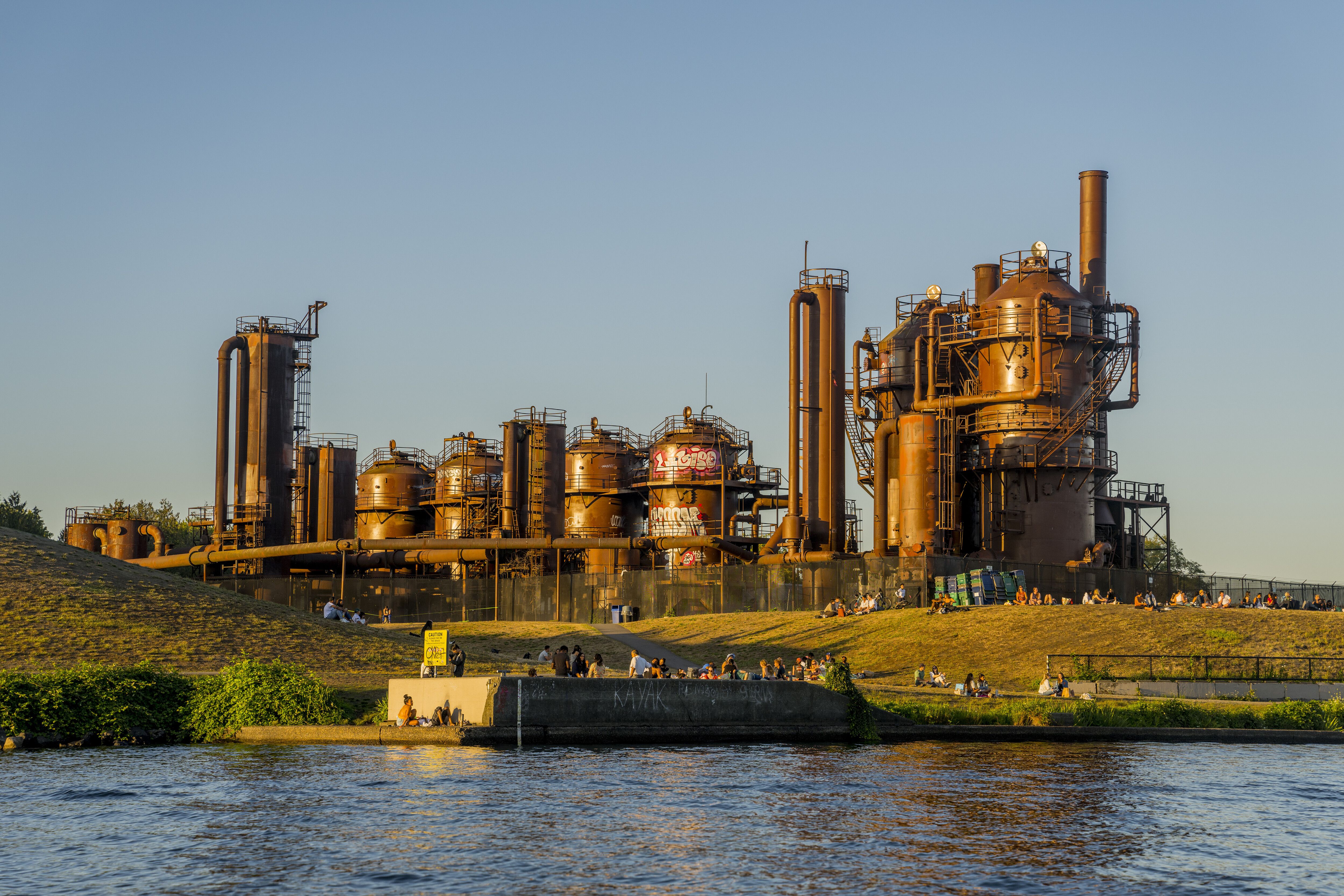 A view of industrial metal towers with round, tall tanks surrounded by ladders, catwalks and platforms, sitting in a grassy area by a lake.