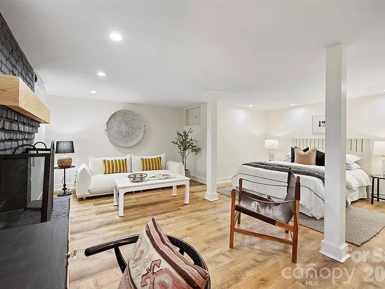 Open basement room with white walls, wood floors, a white sofa with yellow-striped pillows, a bed with gray and beige bedding, dark brick fireplace, and wooden chairs with cushions.