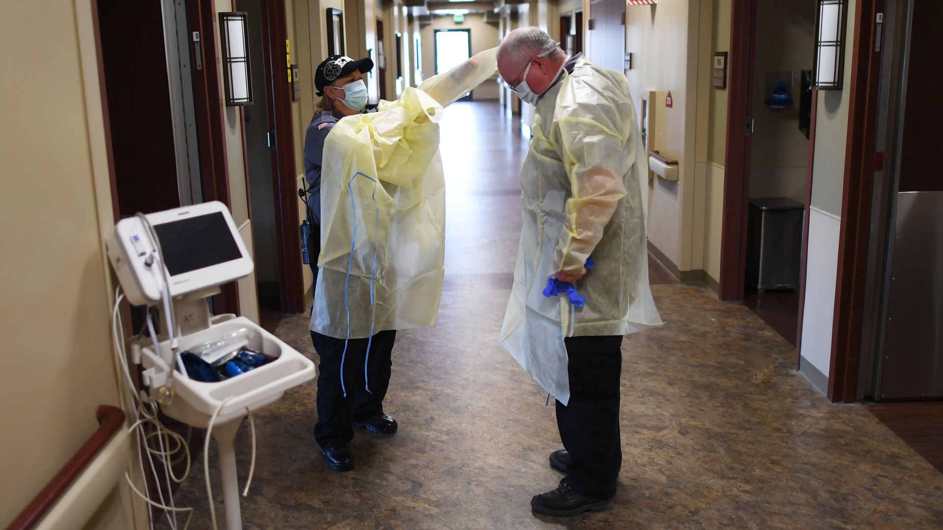 Two EMTs stand in a hospital hallway and put on yellow protective equipment outside a patient rom.