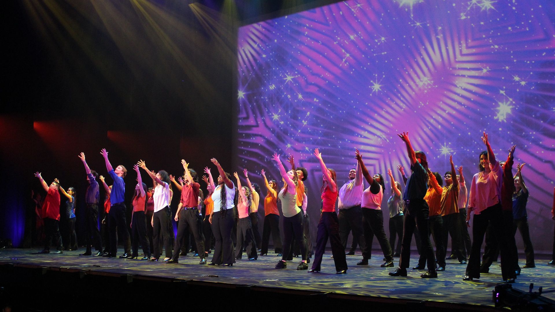 Large group of performers on stage raise their hands in unison; colorful shirts, bright stage lights, and a purple starry backdrop.