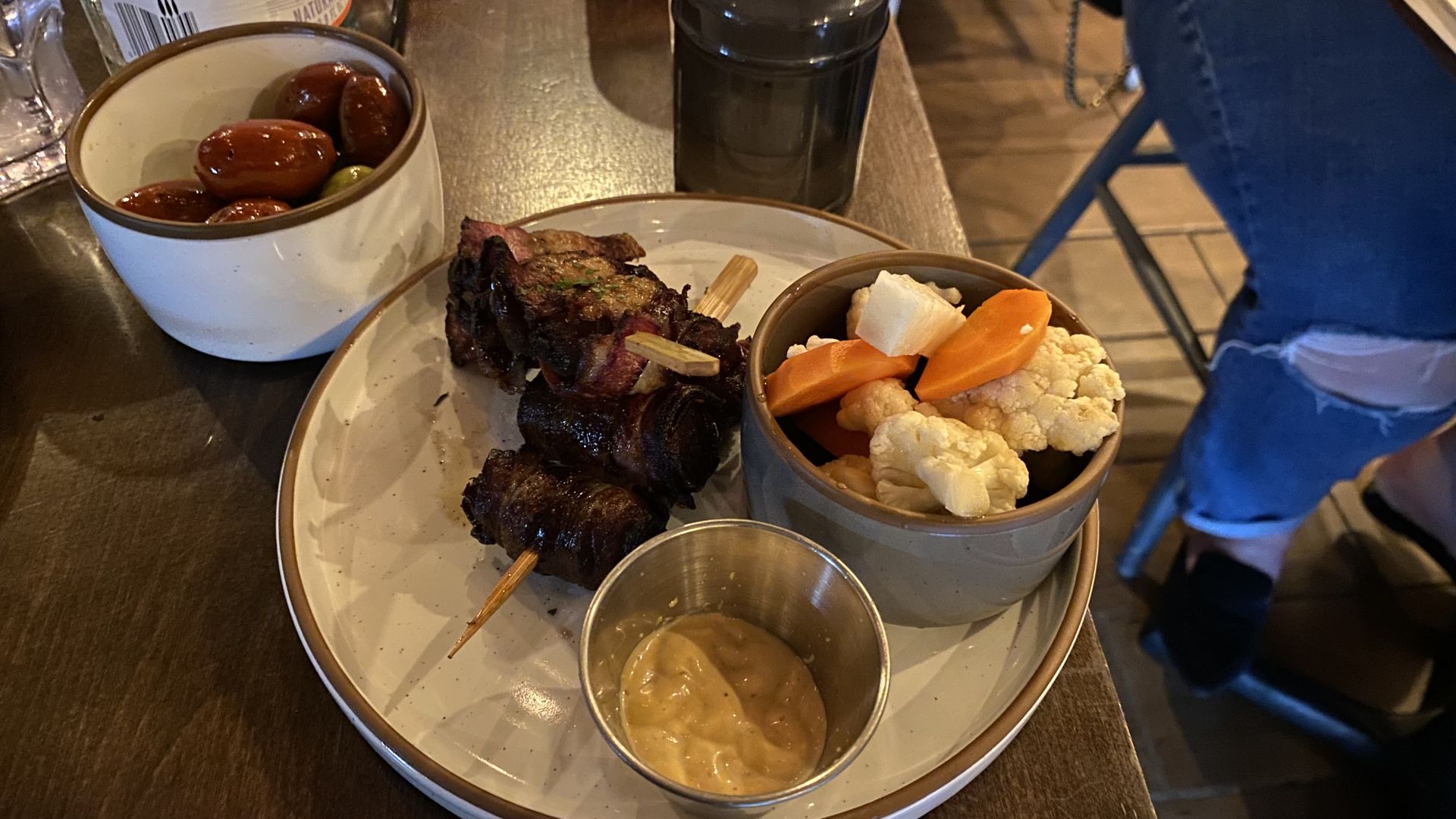 A plate with bacon wrapped dates, sliced carrots and cauliflower and a cup of yellow sauce.
