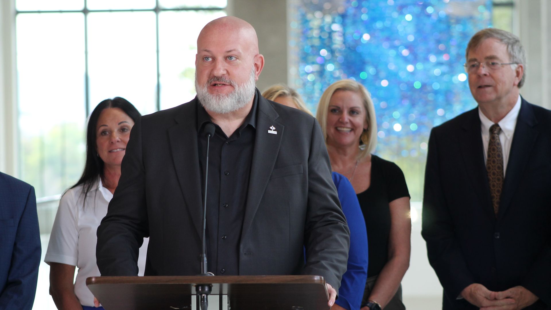 Bearded man in black suit speaking at podium with microphone, flanked by women and a man in business attire, bright window and blue artwork in background.