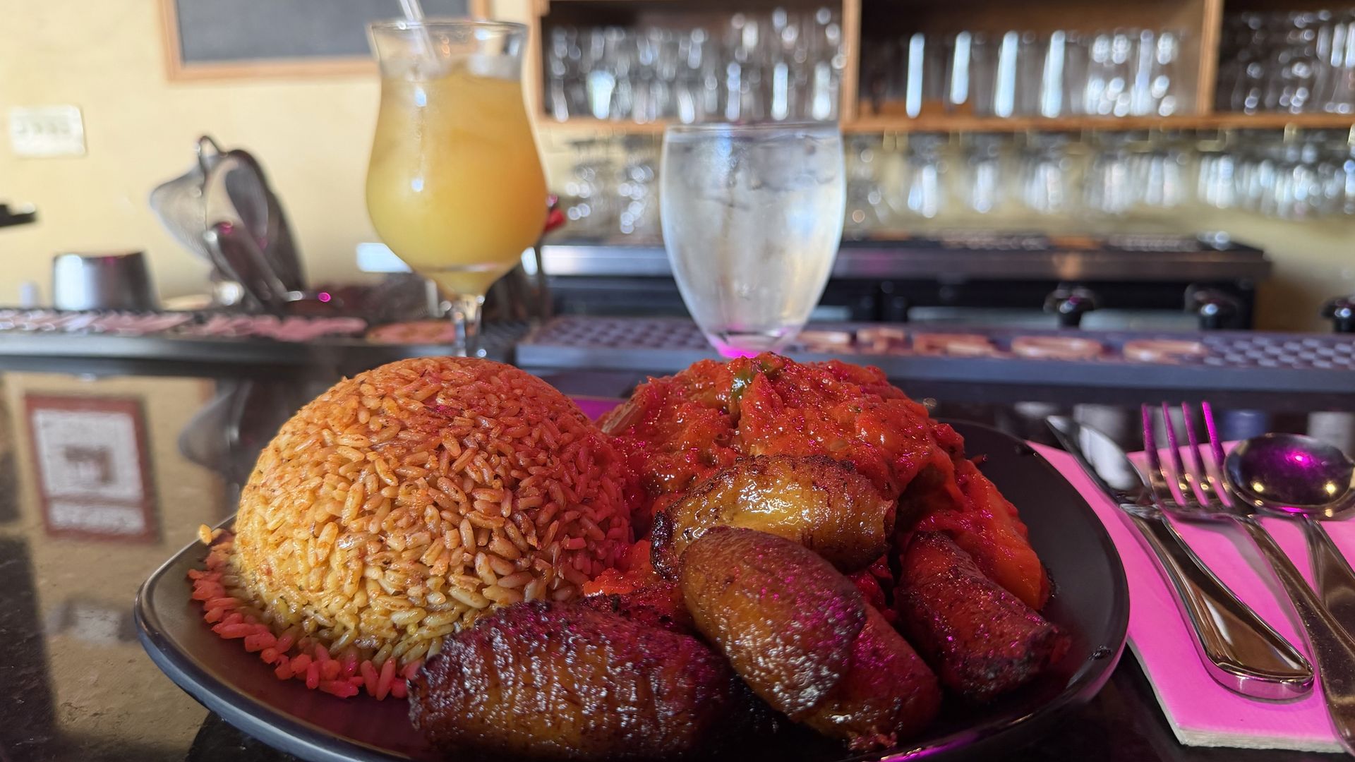 A plate of stewed chicken, jollof rice and plantains at Suya Joint, with a glass of pineapple juice and a glass of water behind the plate.