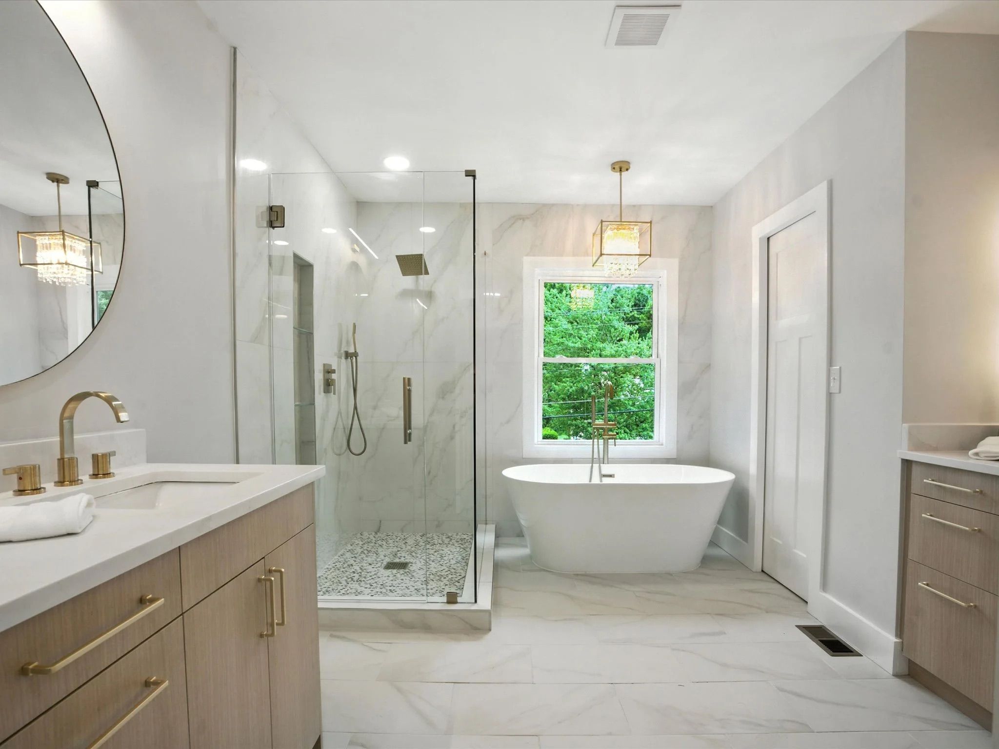 Bright modern bathroom with white freestanding tub by window, glass shower with marble walls, light wood vanity with gold fixtures, and round mirror on pale gray walls.