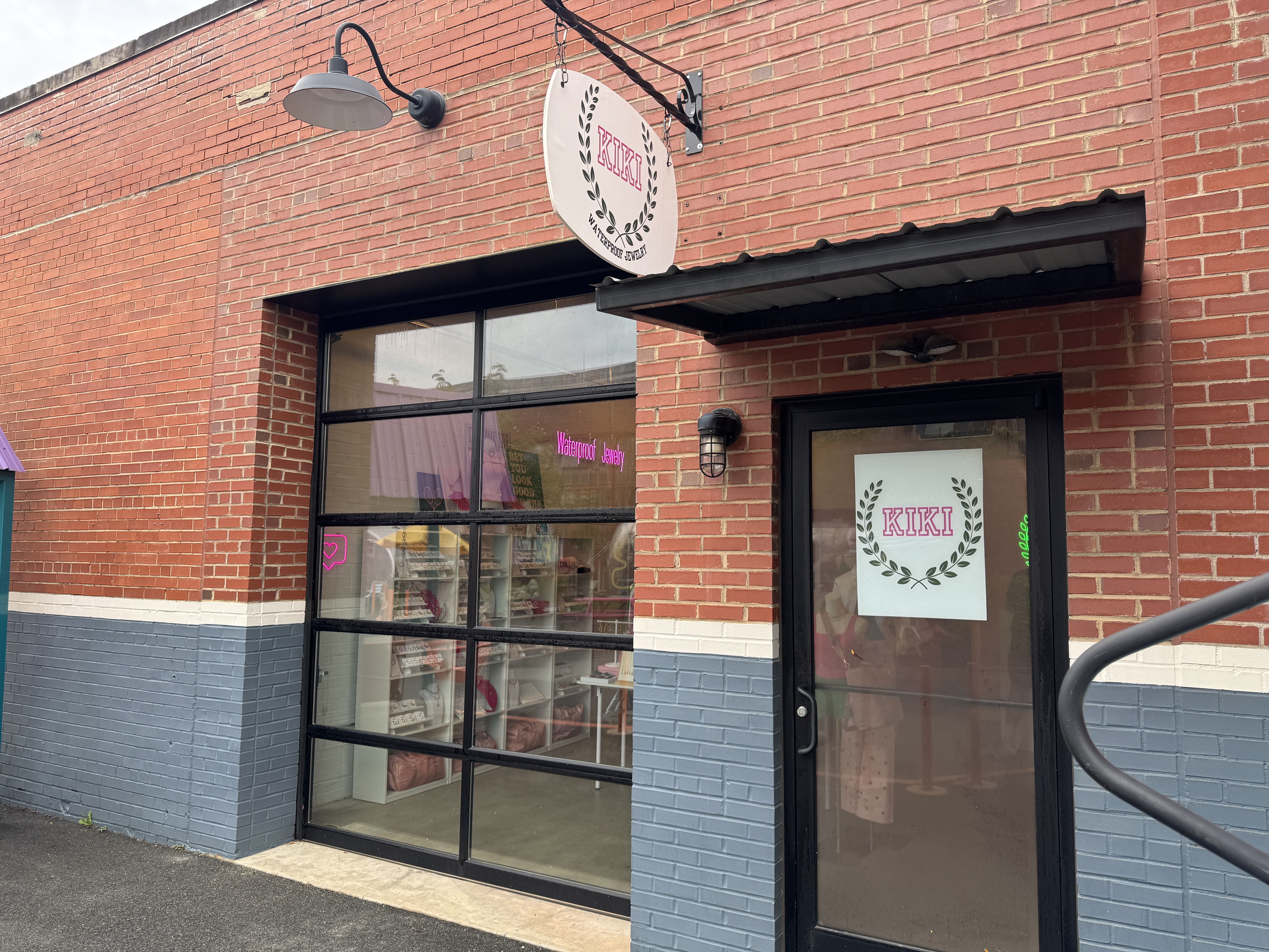 Brick building exterior with black-framed glass door and window. Signs read "KIKI Waterproof Jewelry" with pink neon and printed text. Gray and white painted lower wall.