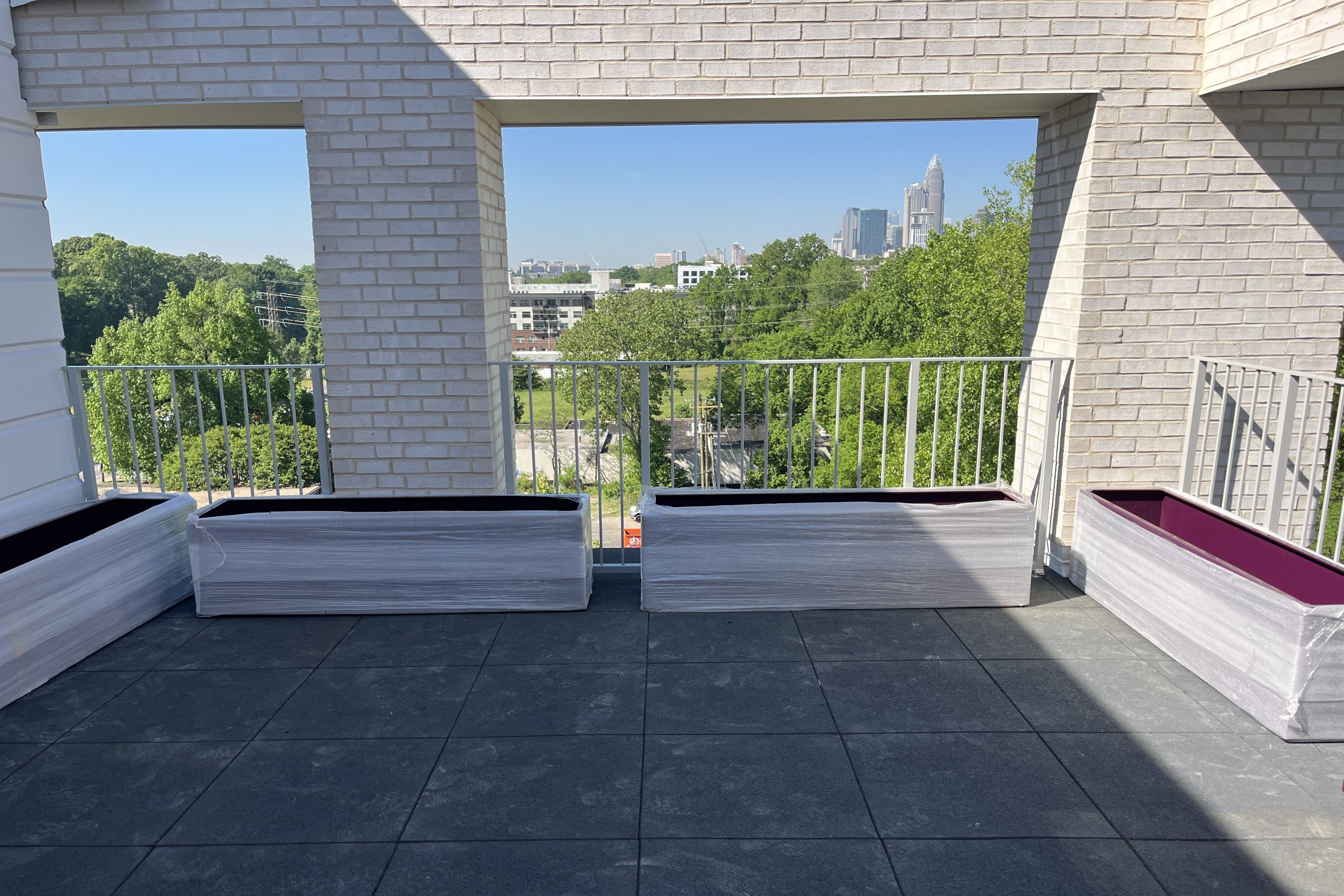 Balcony with white brick columns and metal railing frames a city skyline of tall buildings. In the foreground are wrapped pallets, green trees, and a bright blue sky with a diagonal shadow.