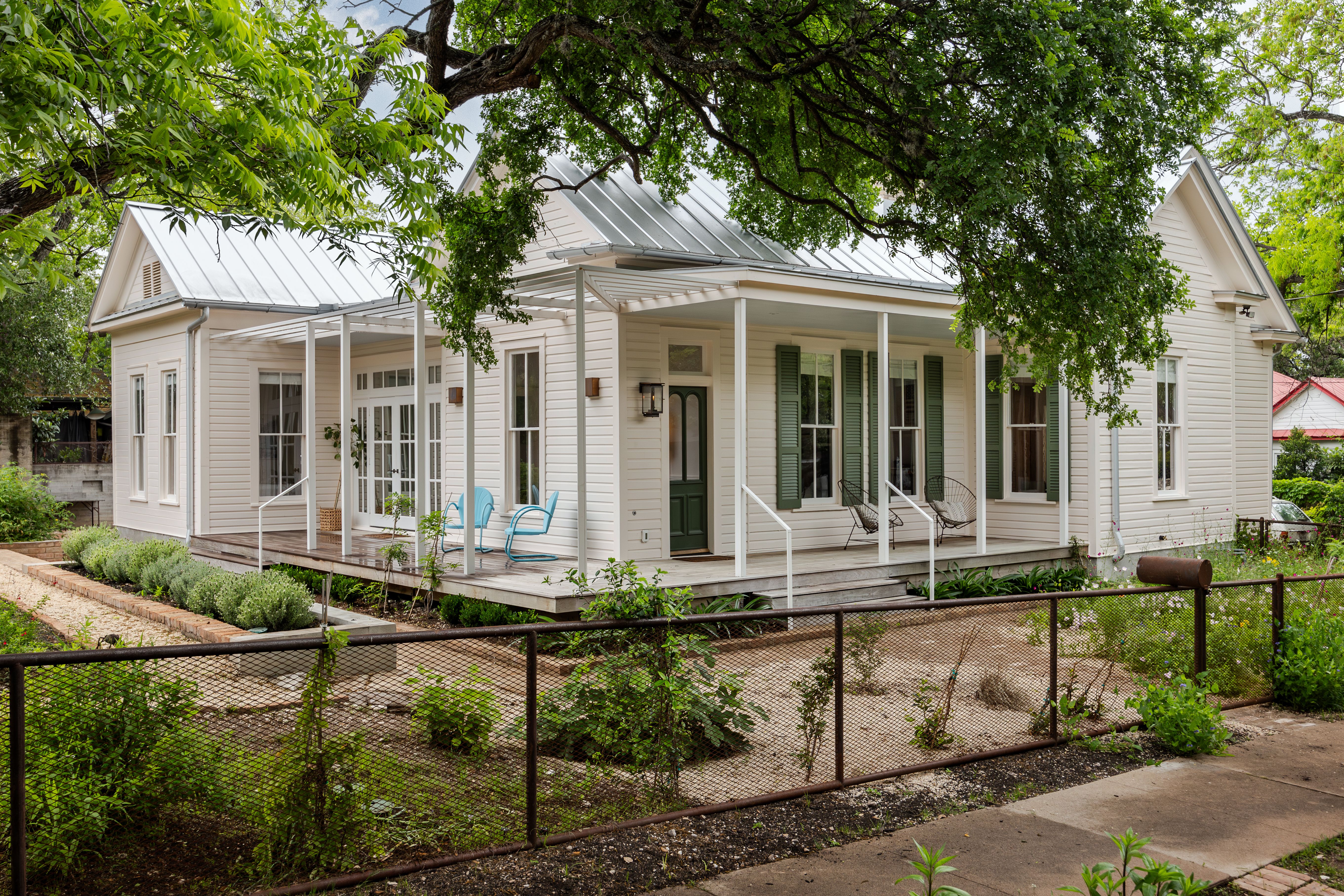 White, single-story cottage with a metal roof and a covered front porch. Green door and shutters, blue chairs on the porch, a brown chain-link fence, and leafy trees framing the yard.