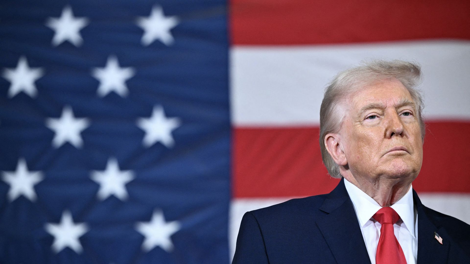 Portrait of President Trump in a navy suit, white shirt, and red tie standing in front of a large American flag with white stars on blue and red and white stripes.
