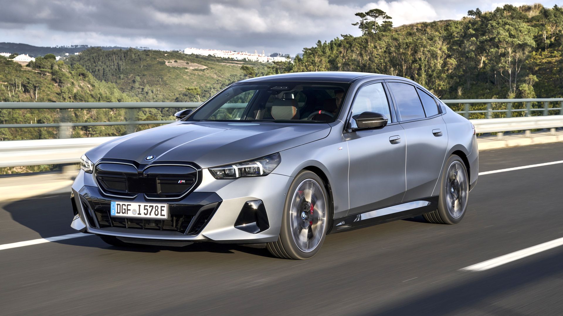 Silver BMW M sedan speeds along a highway bridge with guardrails, set against green hills and a cloudy sky; front grille, headlamps, and large alloy wheels are visible as it passes.