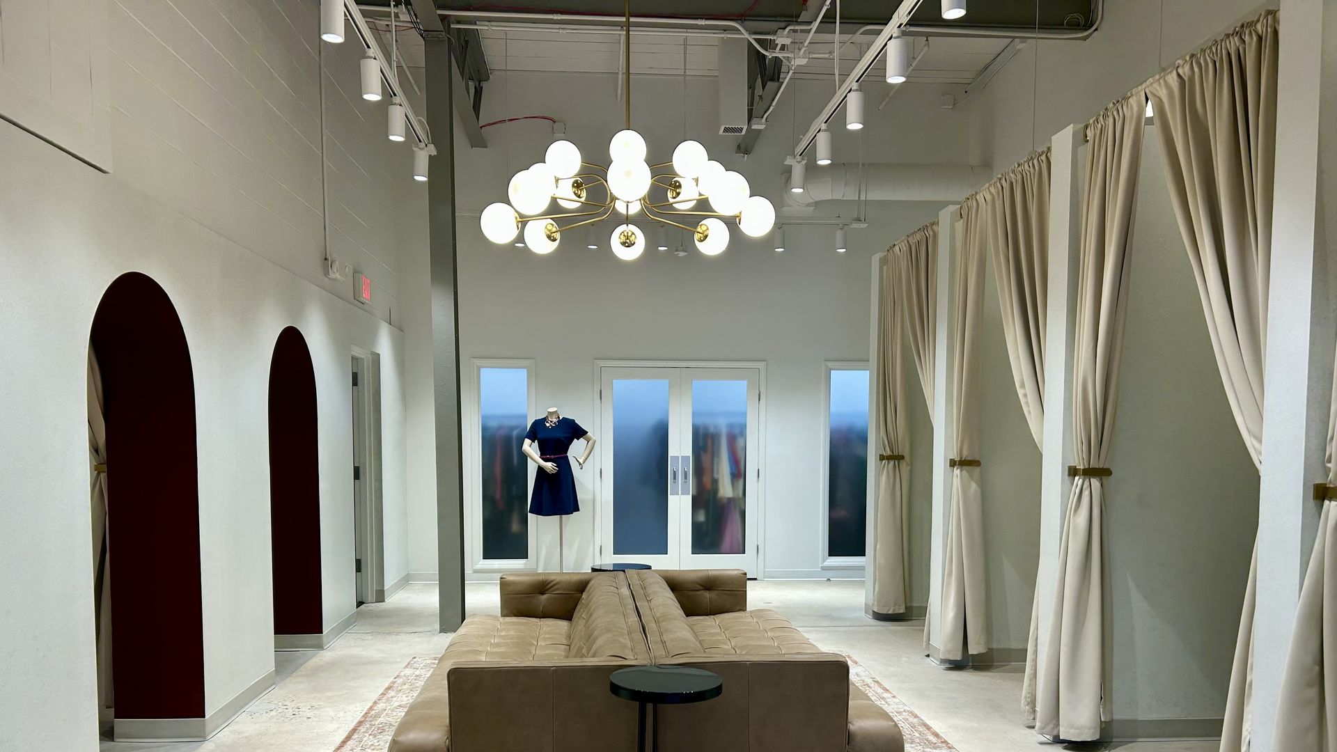 Modern boutique interior with beige leather sofa on patterned rug, round black side table, spherical chandelier, blue dress on mannequin, arched doorways left, and beige dressing room curtains right.