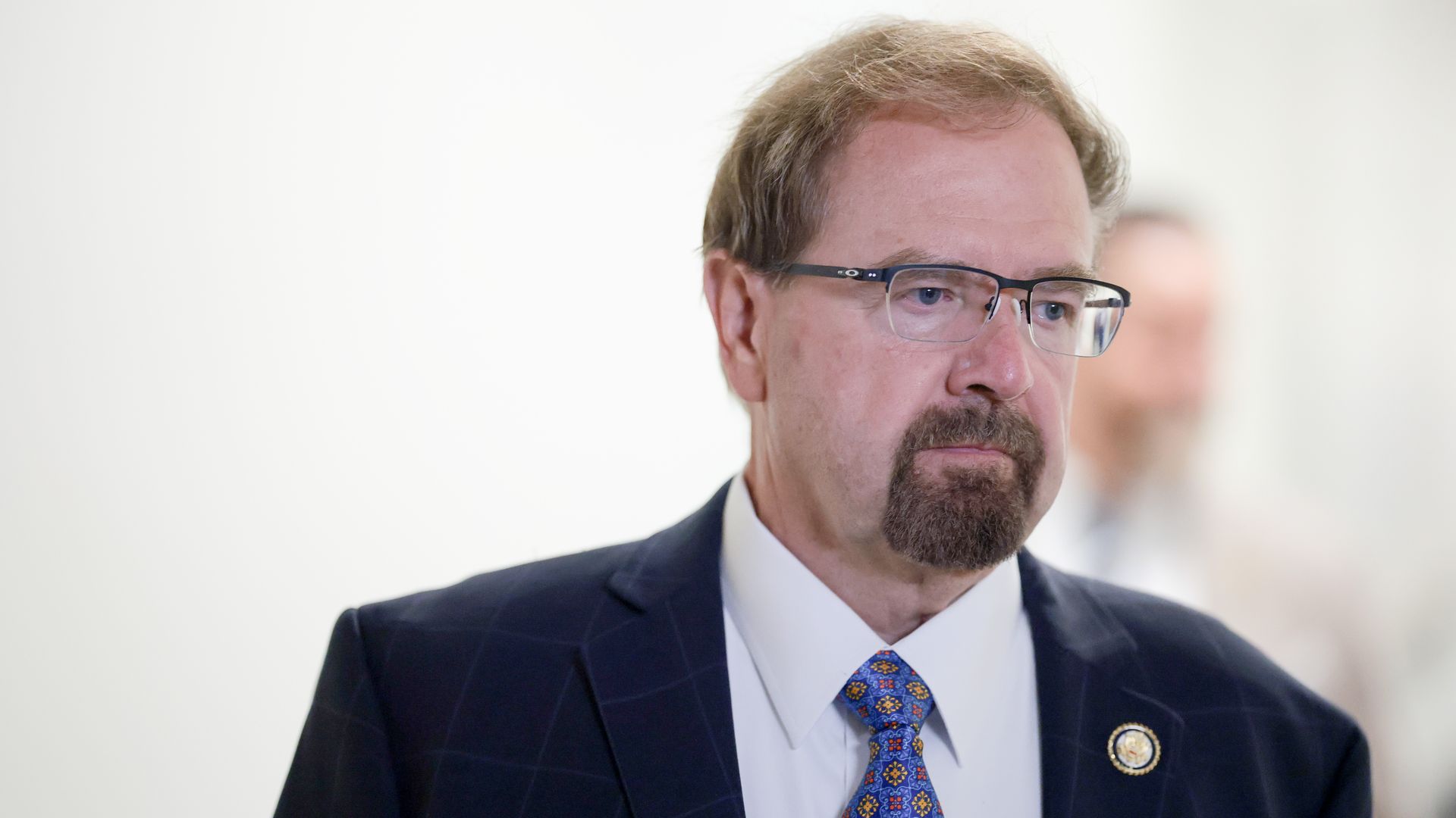 Close-up of a middle-aged man in a dark pinstriped suit, white shirt, and blue patterned tie, wearing black glasses. He has a goatee and light brown hair; a blurred person appears in the background.