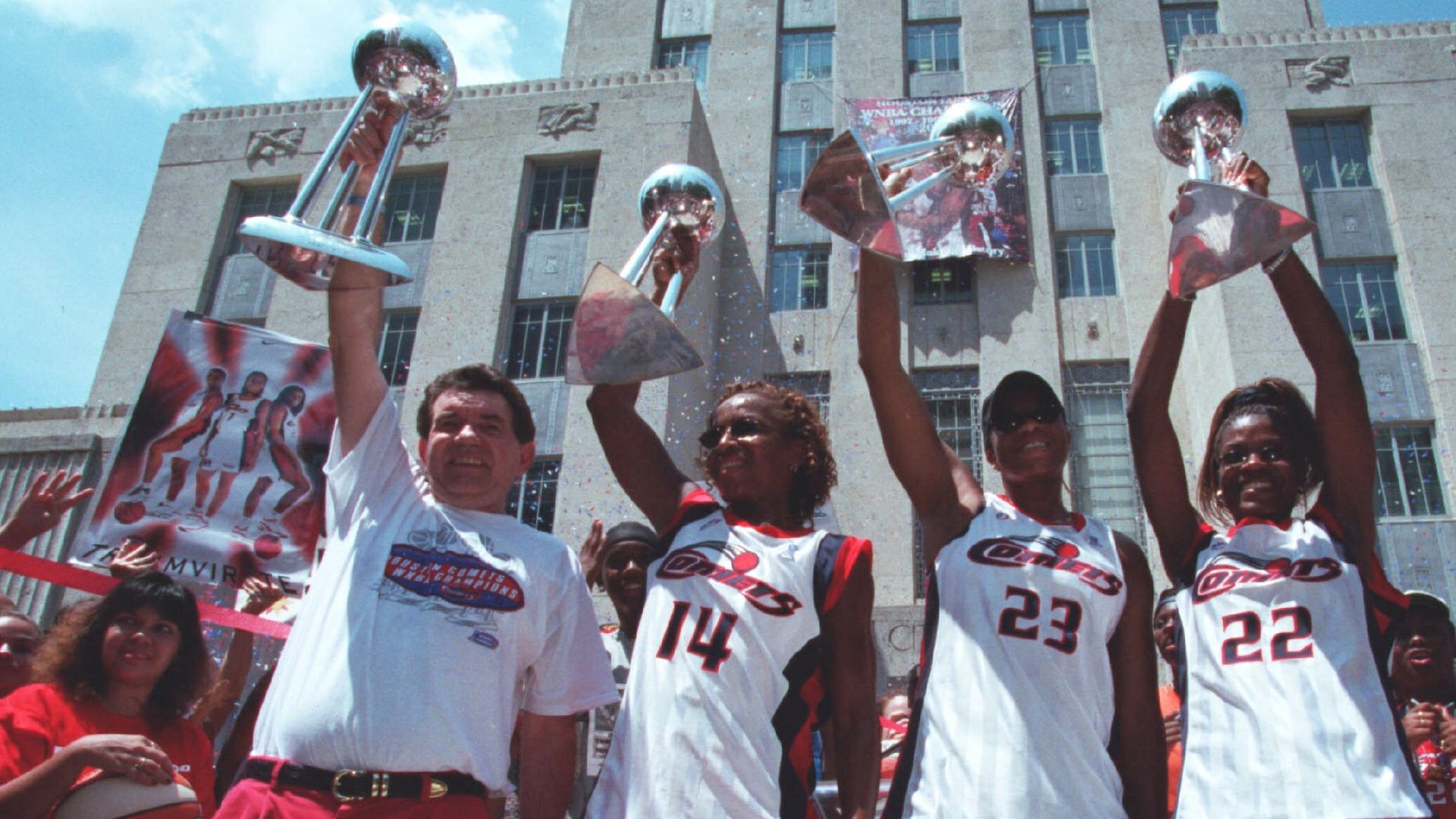 Team members and a coach celebrate outdoors in front of a tall concrete building, raising large silver trophies. They wear white jerseys with red trim and numbers 14, 23, 22; confetti falls.