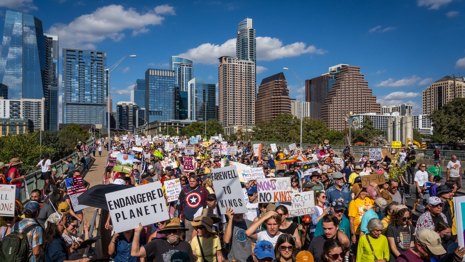Massive crowd of protesters fills a city bridge, holding signs like Endangered Planet and Farewell to Kings, with a skyline of glass towers beneath a bright blue sky.