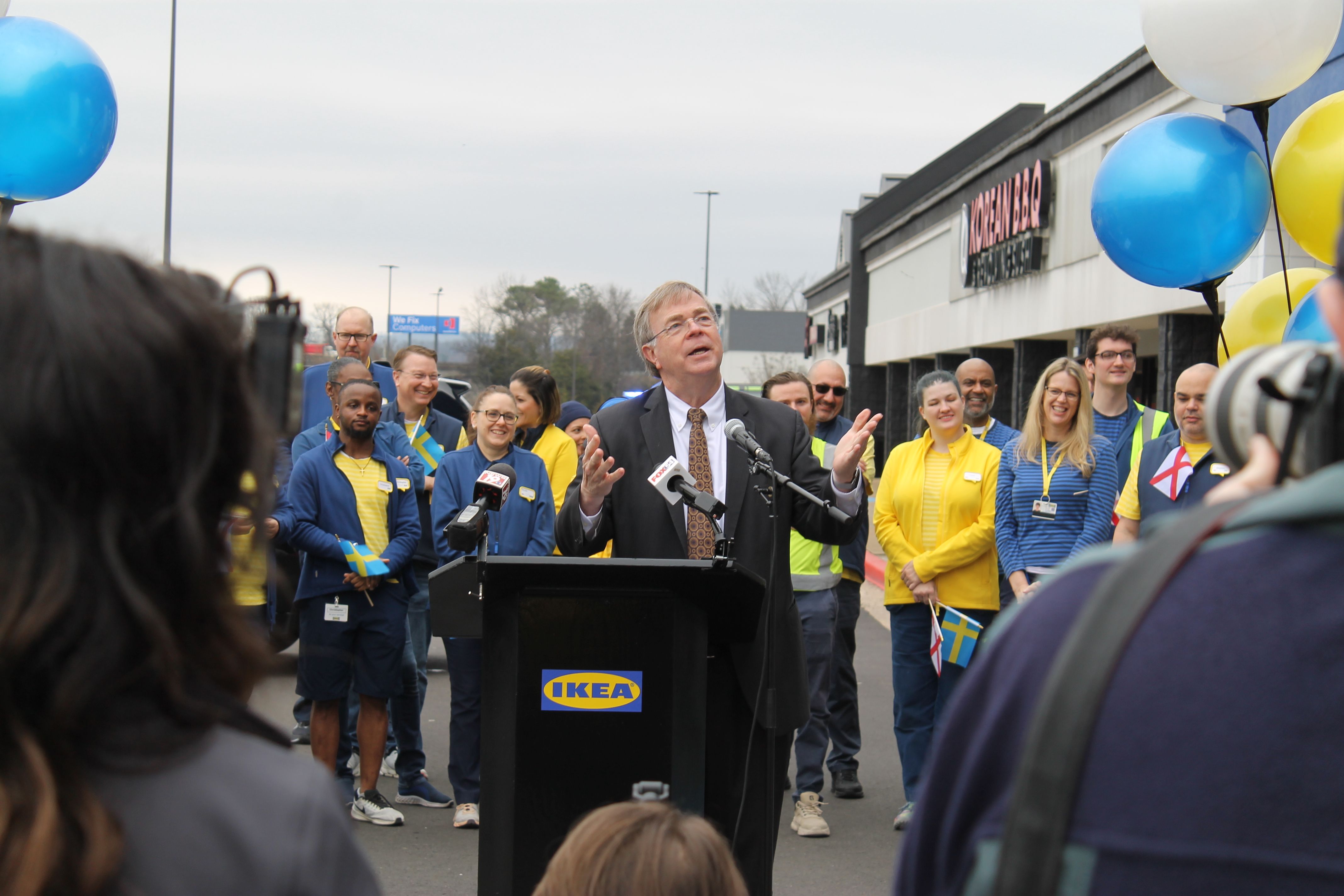 A man in a black suit speaks at a podium with an IKEA logo, surrounded by smiling people in blue and yellow clothing holding Swedish flags and balloons, outside a shopping mall.