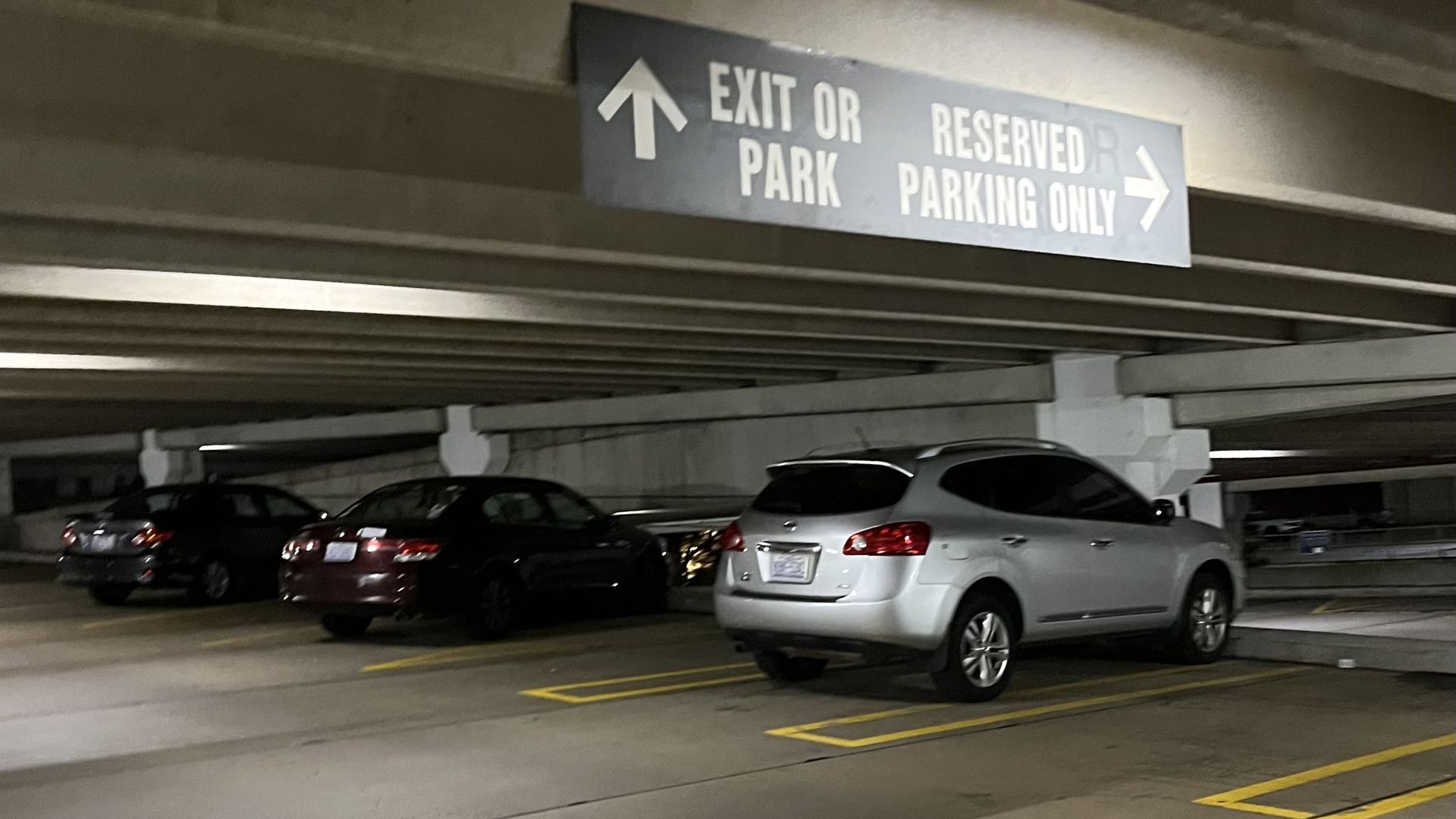 Indoor parking garage with a blue ceiling sign reading EXIT OR PARK with a left arrow and RESERVED PARKING ONLY with a right arrow; several cars parked below.