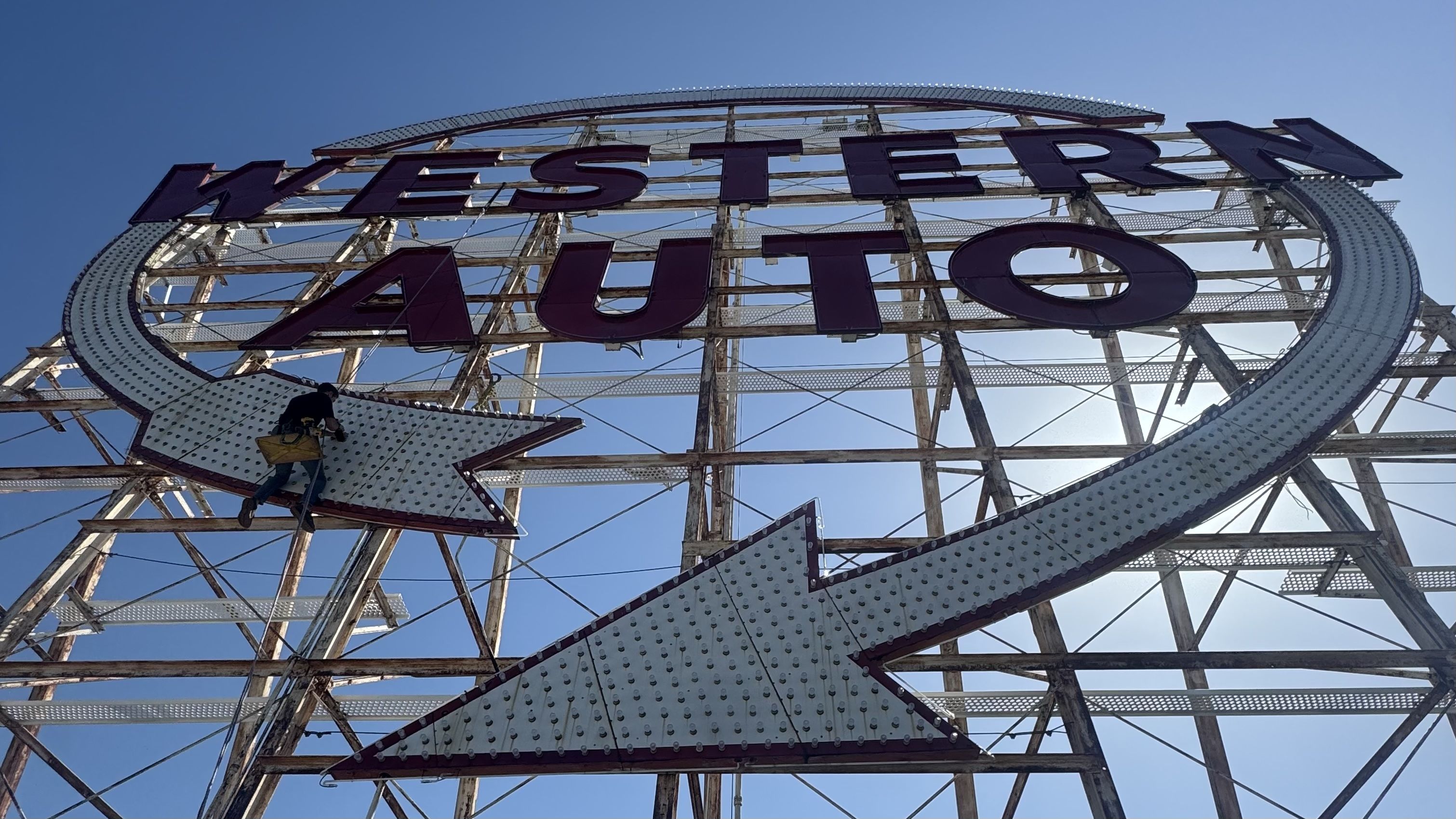 Worker on a suspended platform repairs a large vintage WESTERN AUTO sign with bold purple letters inside a circular dotted border atop a rusted metal framework against a blue sky.