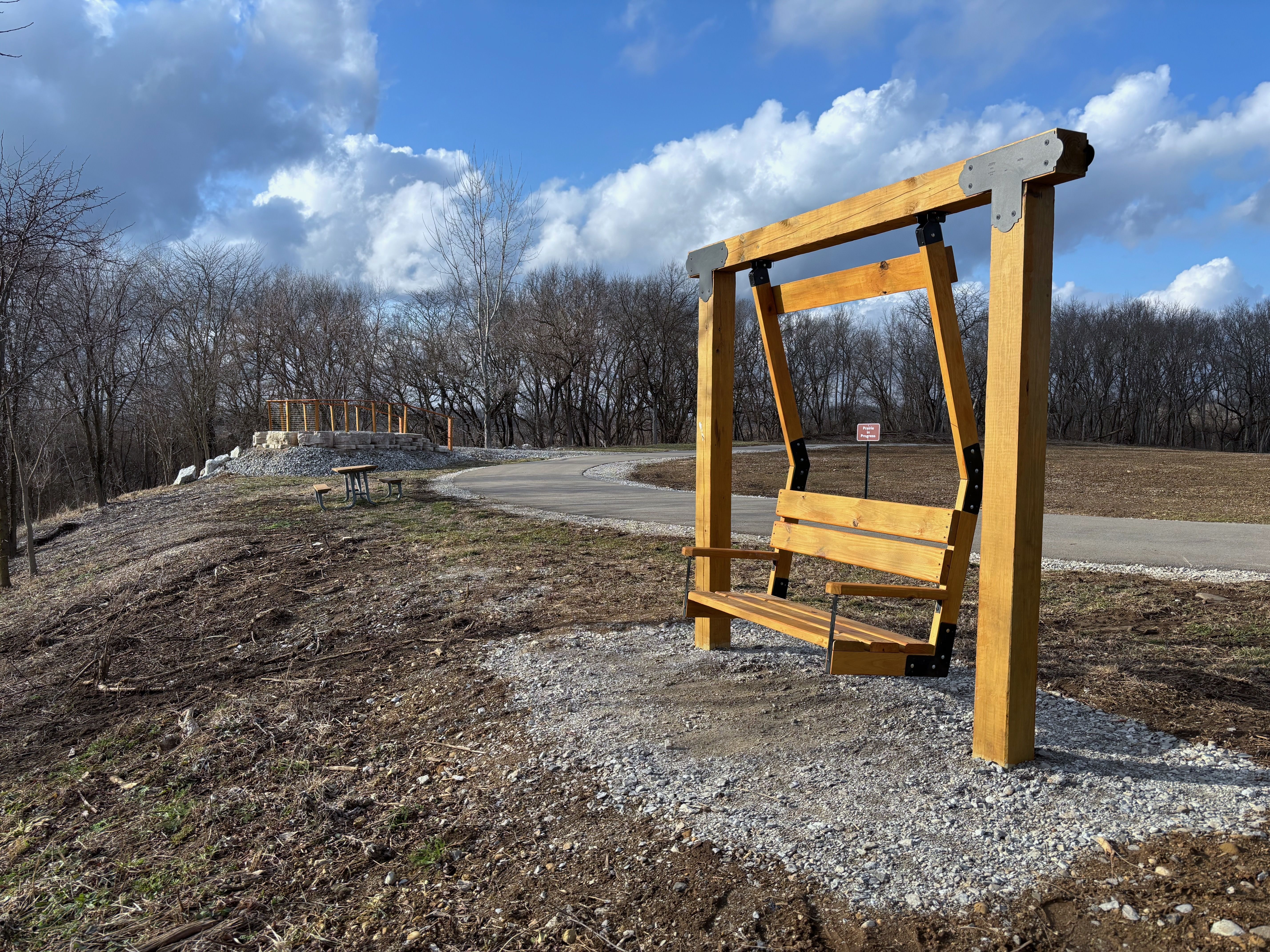 Wooden bench swing with metal brackets on gravel by a paved path in a park area with leafless trees, picnic table, and partly cloudy blue sky.