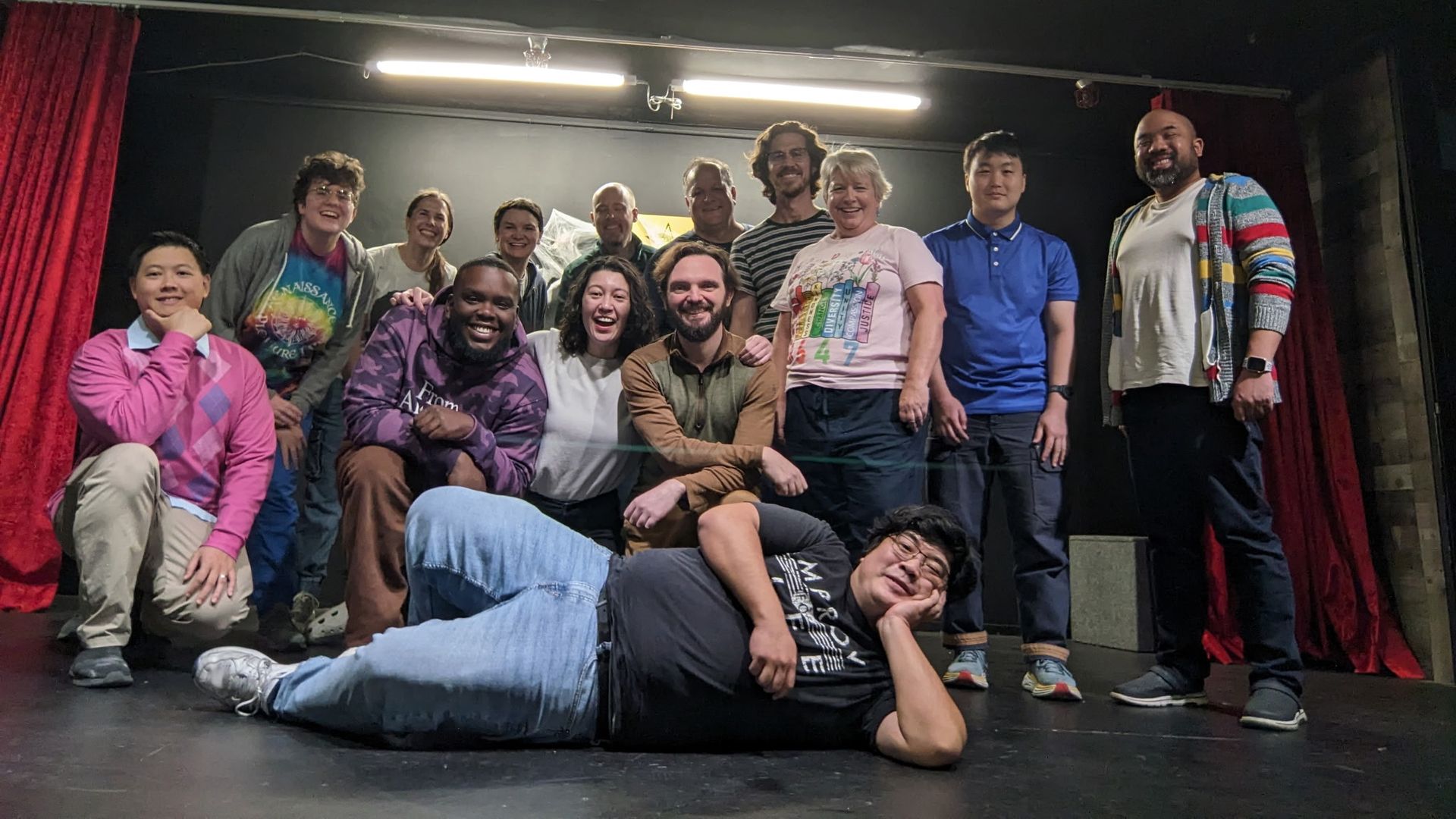 Group of 14 diverse people posing happily on a stage with red curtains and black walls; one person lies in front wearing glasses and a black shirt, others stand or kneel behind.