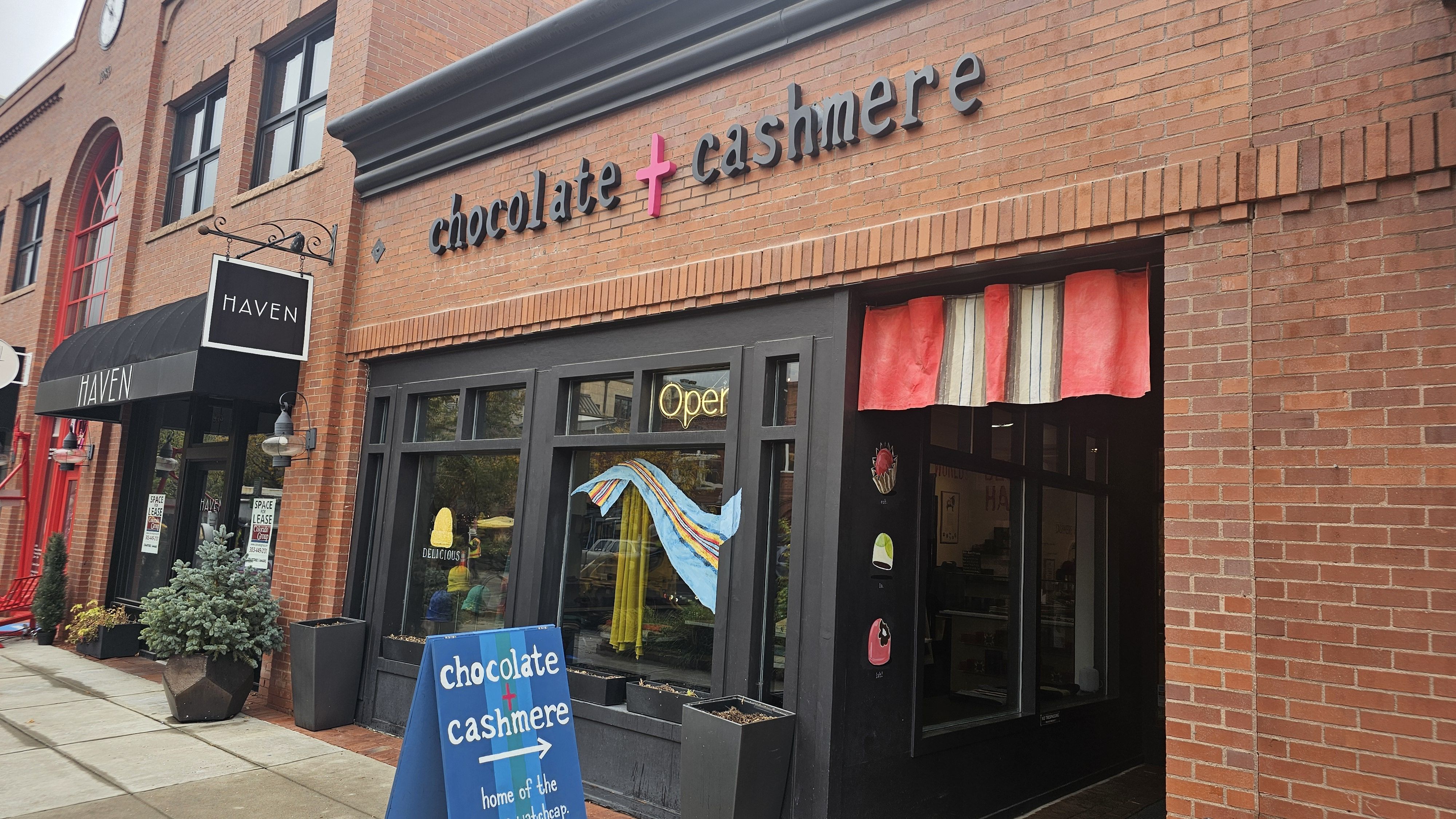 Brick storefront with a large 'chocolate + cashmere' sign and pink cross. Black-framed windows and a black 'HAVEN' awning; red/cream striped curtains inside. A blue sign on the sidewalk reads 'chocolate cashmere'.