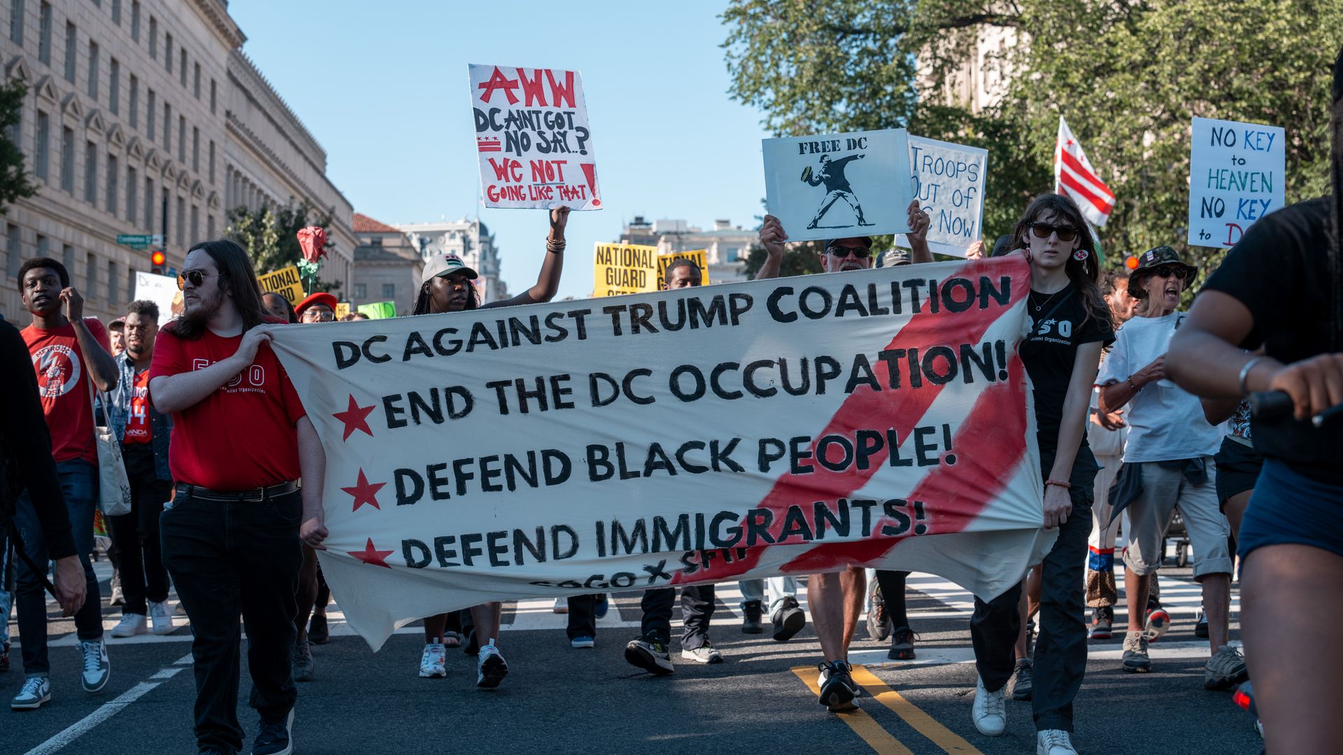 Protesters carry signs opposing Trump's takeover of D.C. with phrases like "DC AGAINST TRUMP COALITION" as they march.