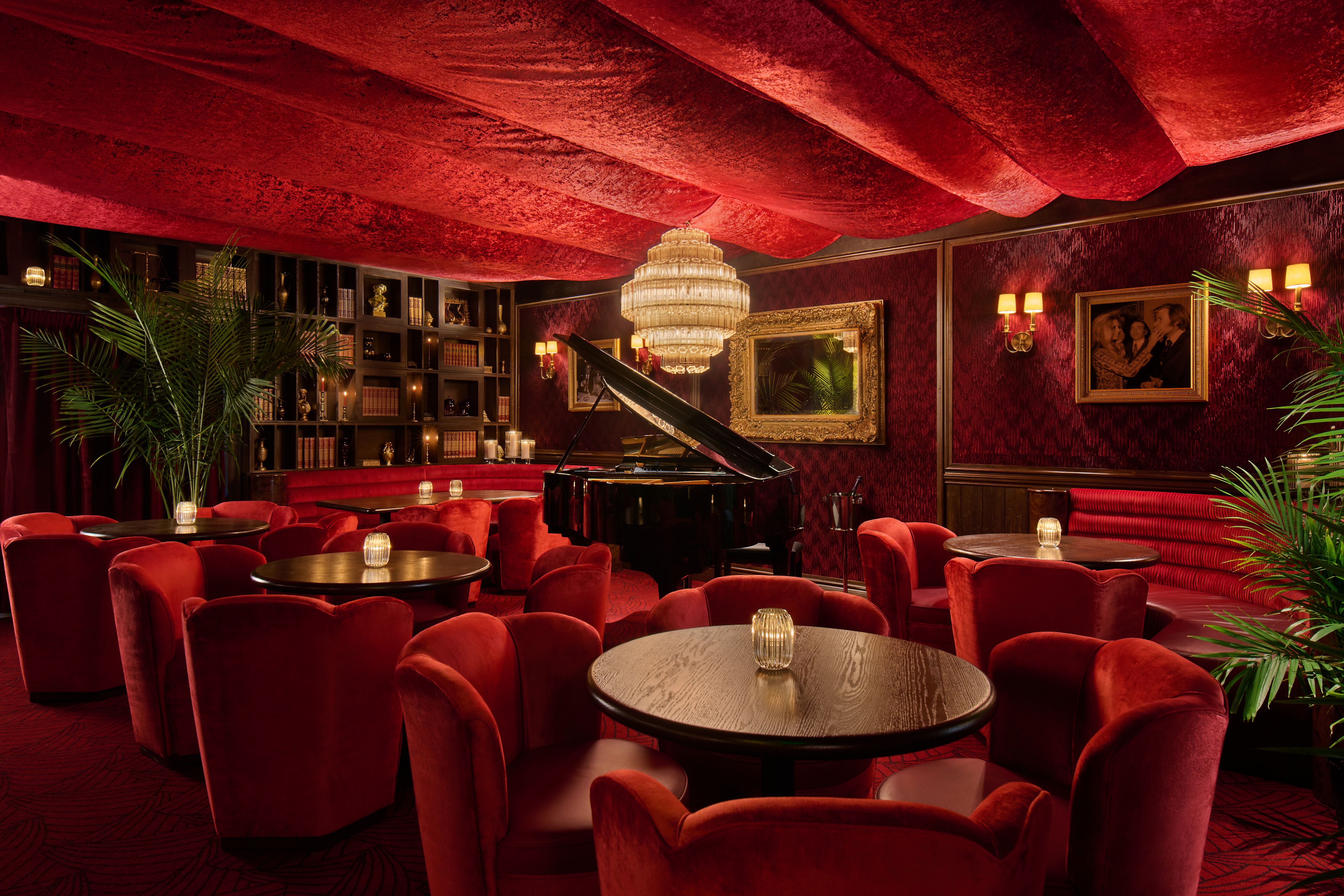 Luxurious red lounge with velvet chairs around round tables, a grand piano, crystal chandelier, red draped ceiling, gold-framed mirror, dark wood bookshelves, warm wall sconces, and green plants.