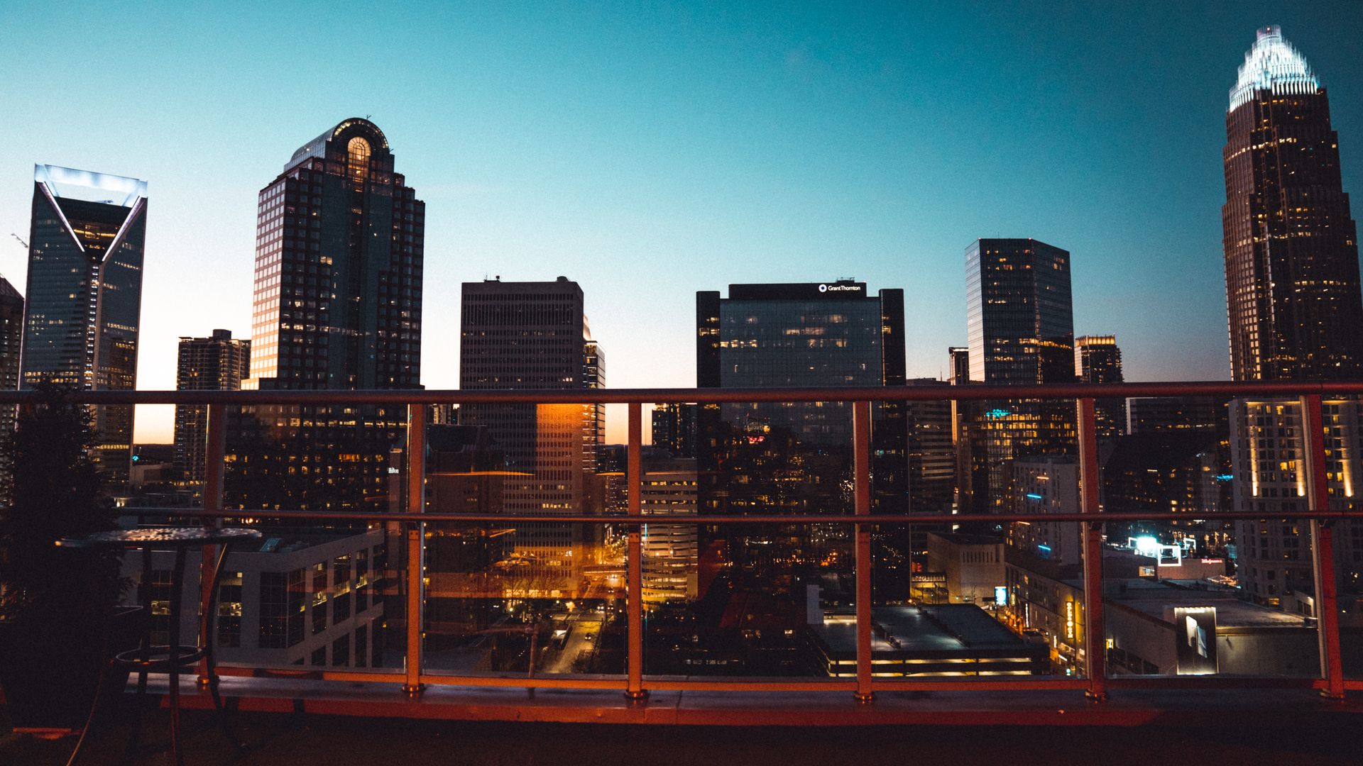 Rooftop cityscape at dusk: a railing in the foreground, tall illuminated skyscrapers, and a blue-to-orange gradient sky with lights glowing below.